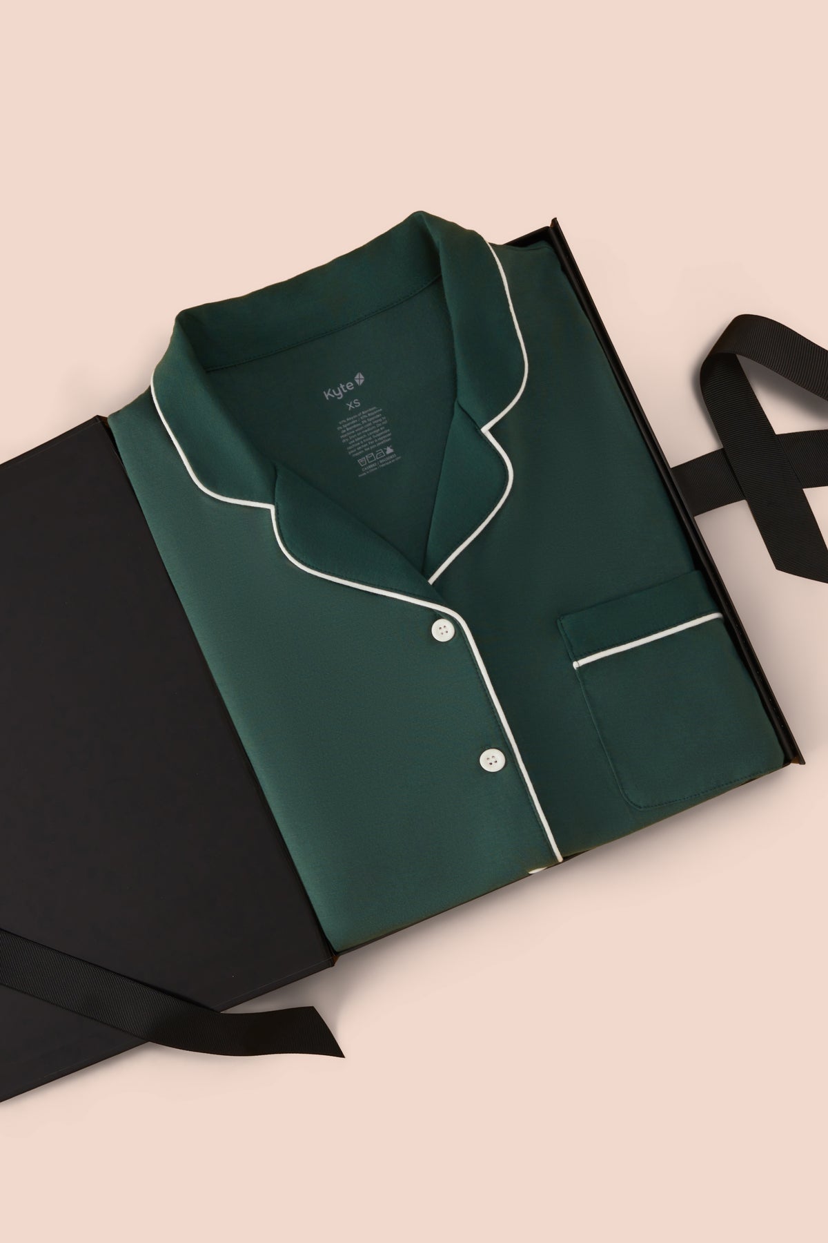 A folded dark green pajama shirt with white piping, presented in a black gift box on a light beige background.