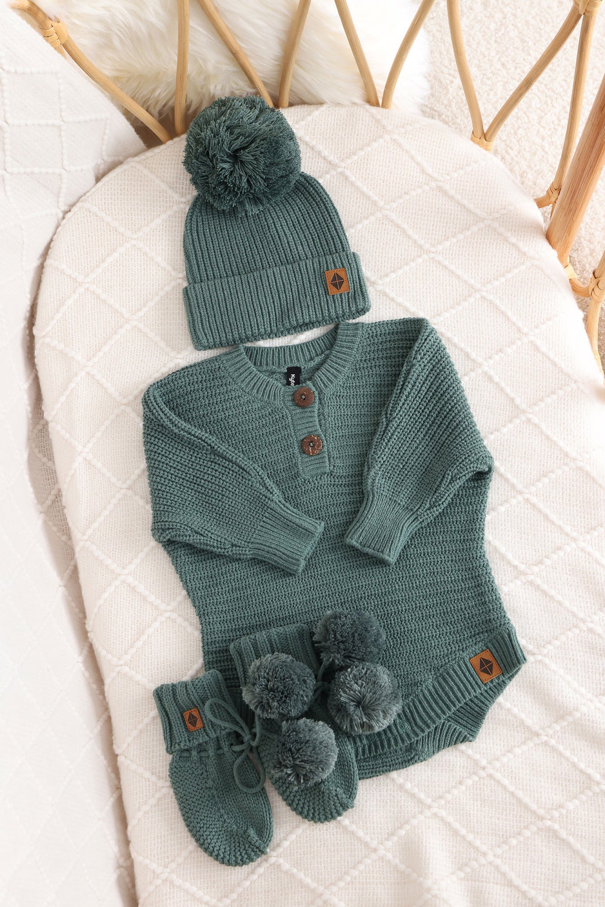 Chunky knit pom beanie, chunky knit romper and knit booties in pine laid in a crib