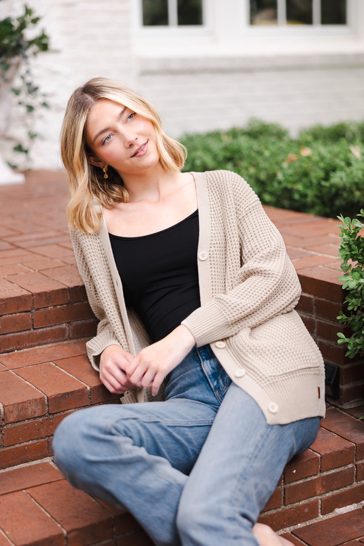 Modal sitting outdoors wearing Chunky Knit Women's Waffle Cardigan in Almond over black camisole and blue jeans