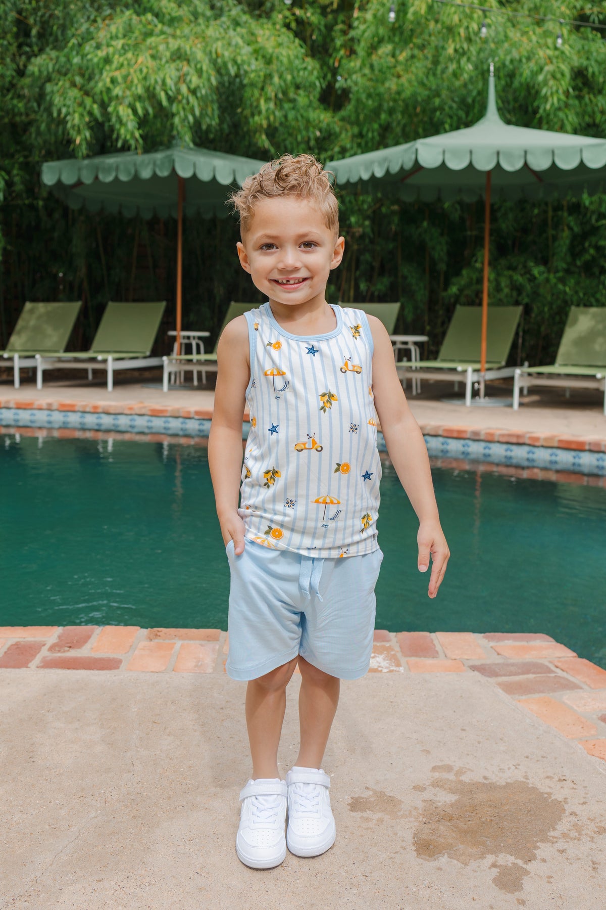 Young boy wearing a tank top with amalfi coast inspired print and blue shorts by a pool