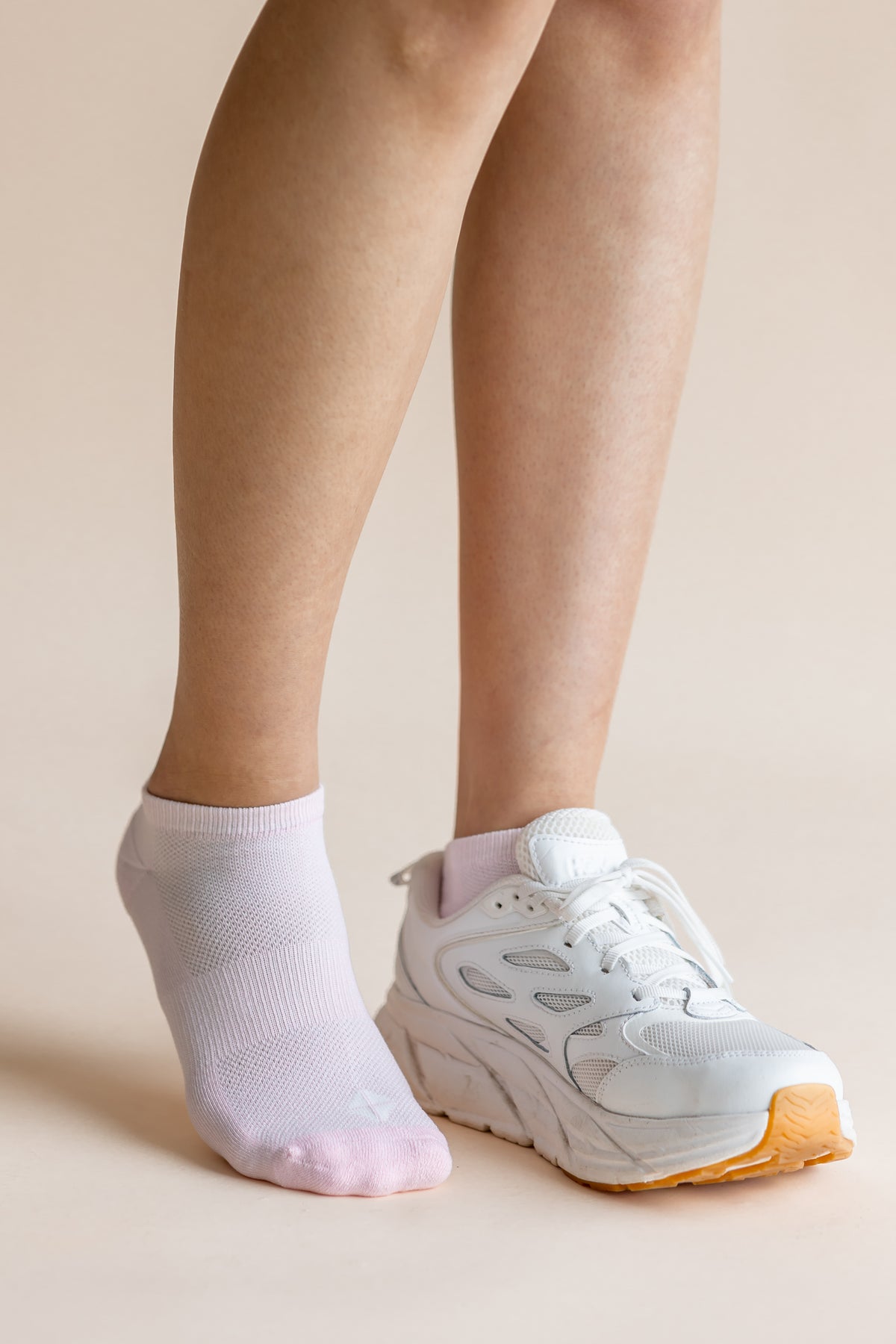 A person's legs and feet shown wearing a pair of white ankle socks, with one foot in a white sneaker and the other barefoot, against a solid beige background.