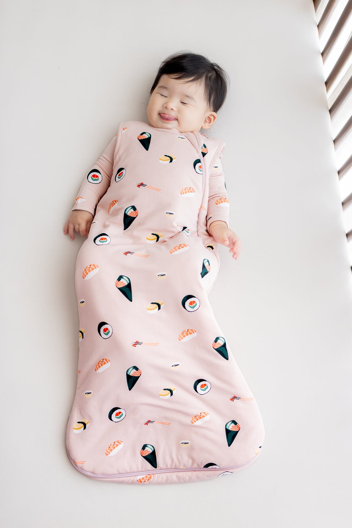 Baby laying in crib with white sheets in blush sushi sleep bag
