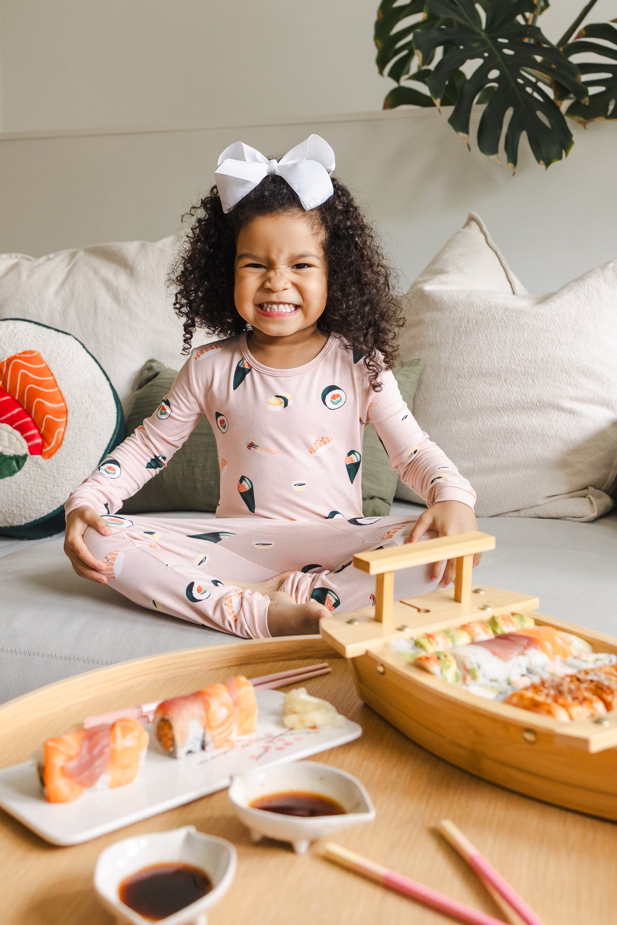 Child sitting crisscross on a white couch wearing blush sushi pajama set and white bow with sushi on a table in front of her