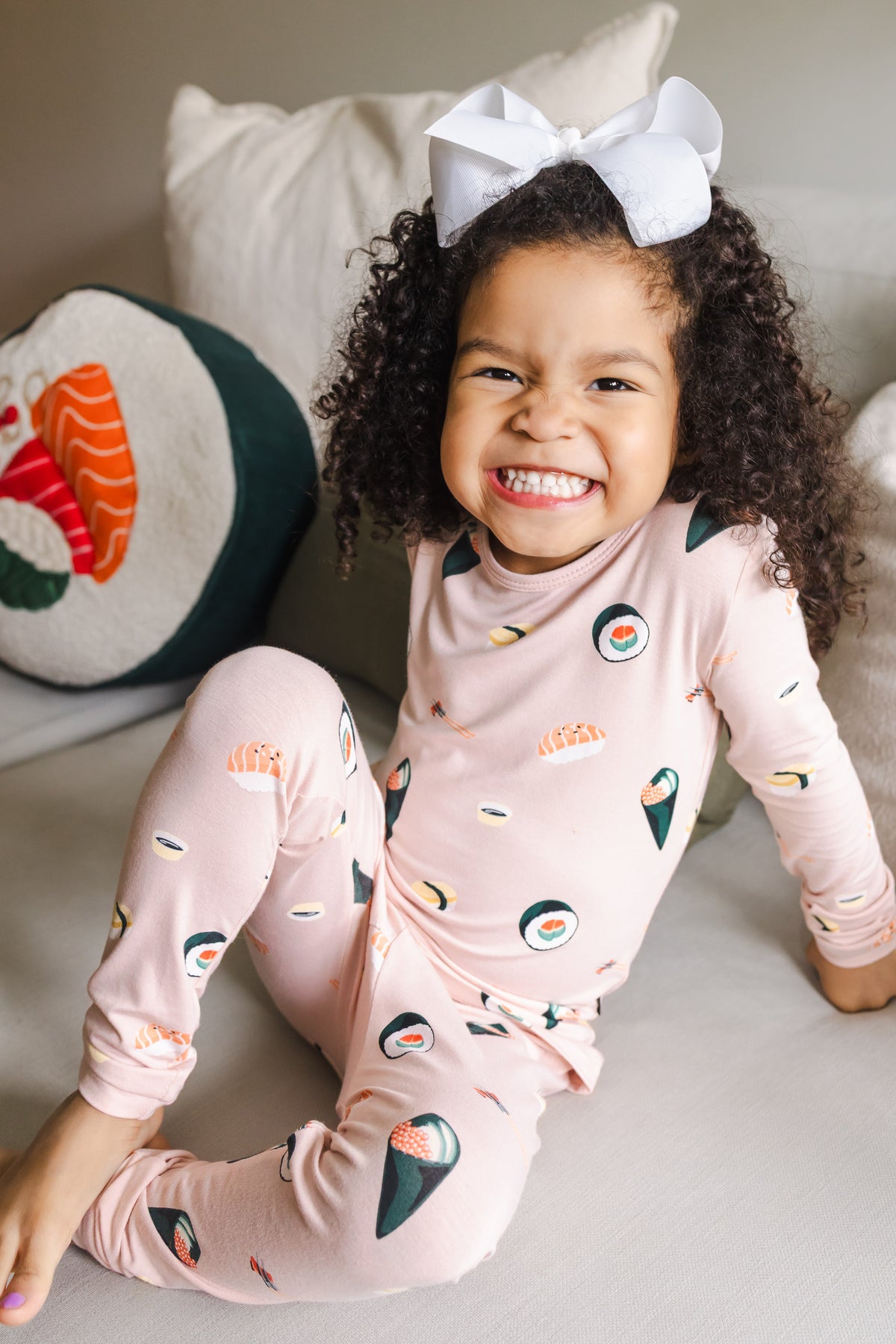 Child smiling sitting on a white couch with one leg up wearing blush sushi pajama set with a white bow