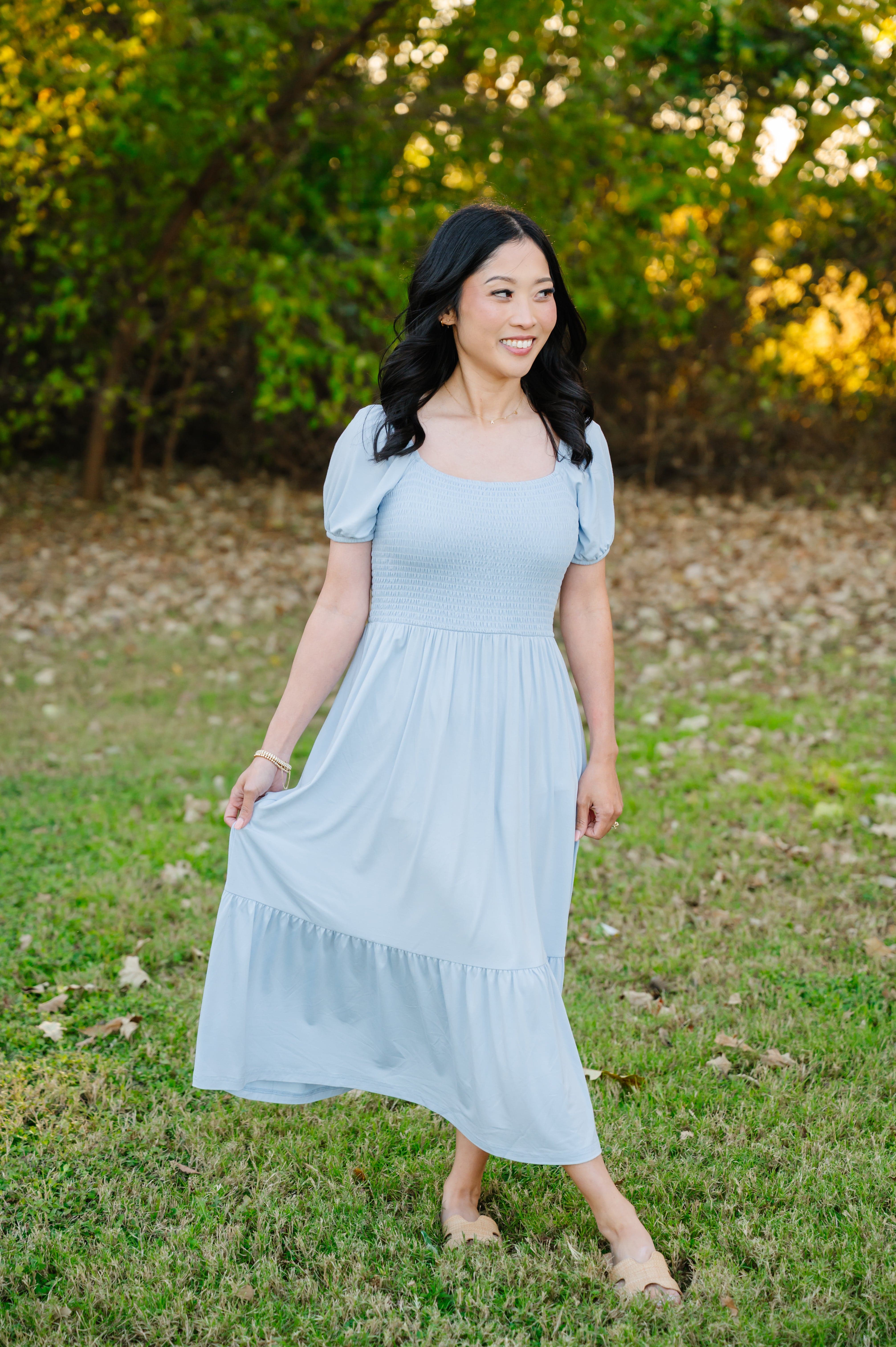 Female modeling the Women’s Smocked Dress in Breeze standing in a grassy field surrounded by trees
