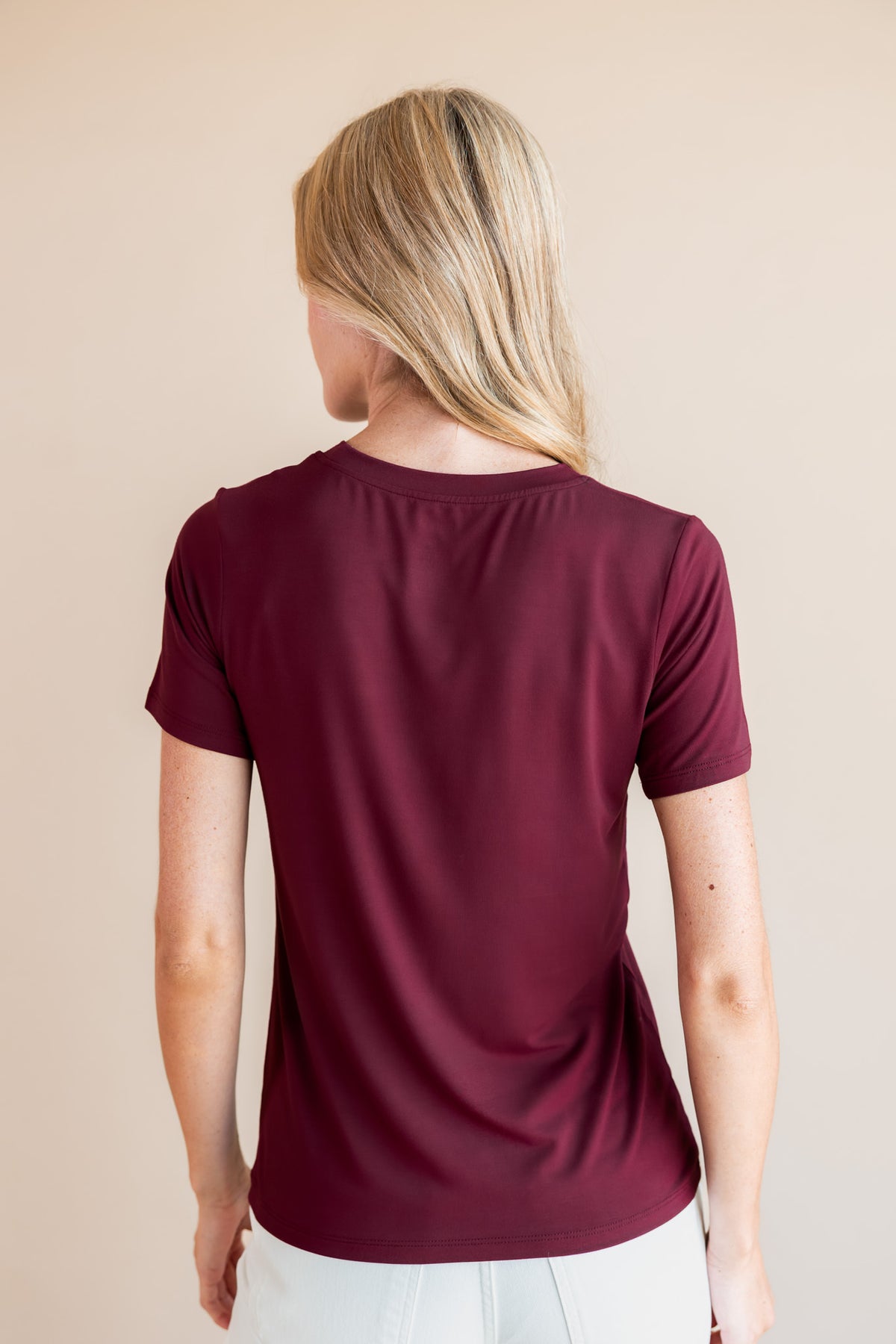 A woman with blonde hair is shown from the back wearing a short-sleeve crewneck t-shirt in burgundy, paired with white pants, against a solid beige background.