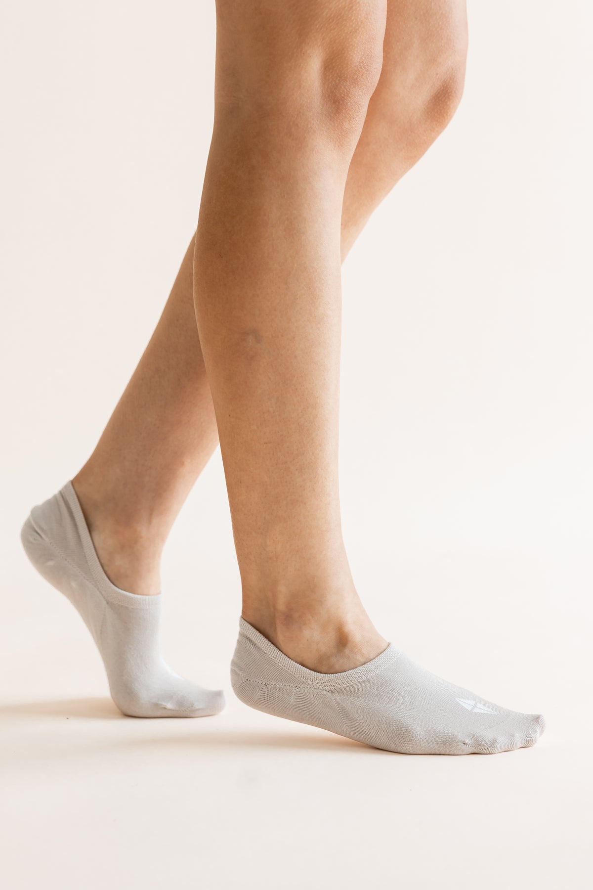 A close-up of a person's feet wearing light gray low-cut no-show socks against a plain, warm off-white background.