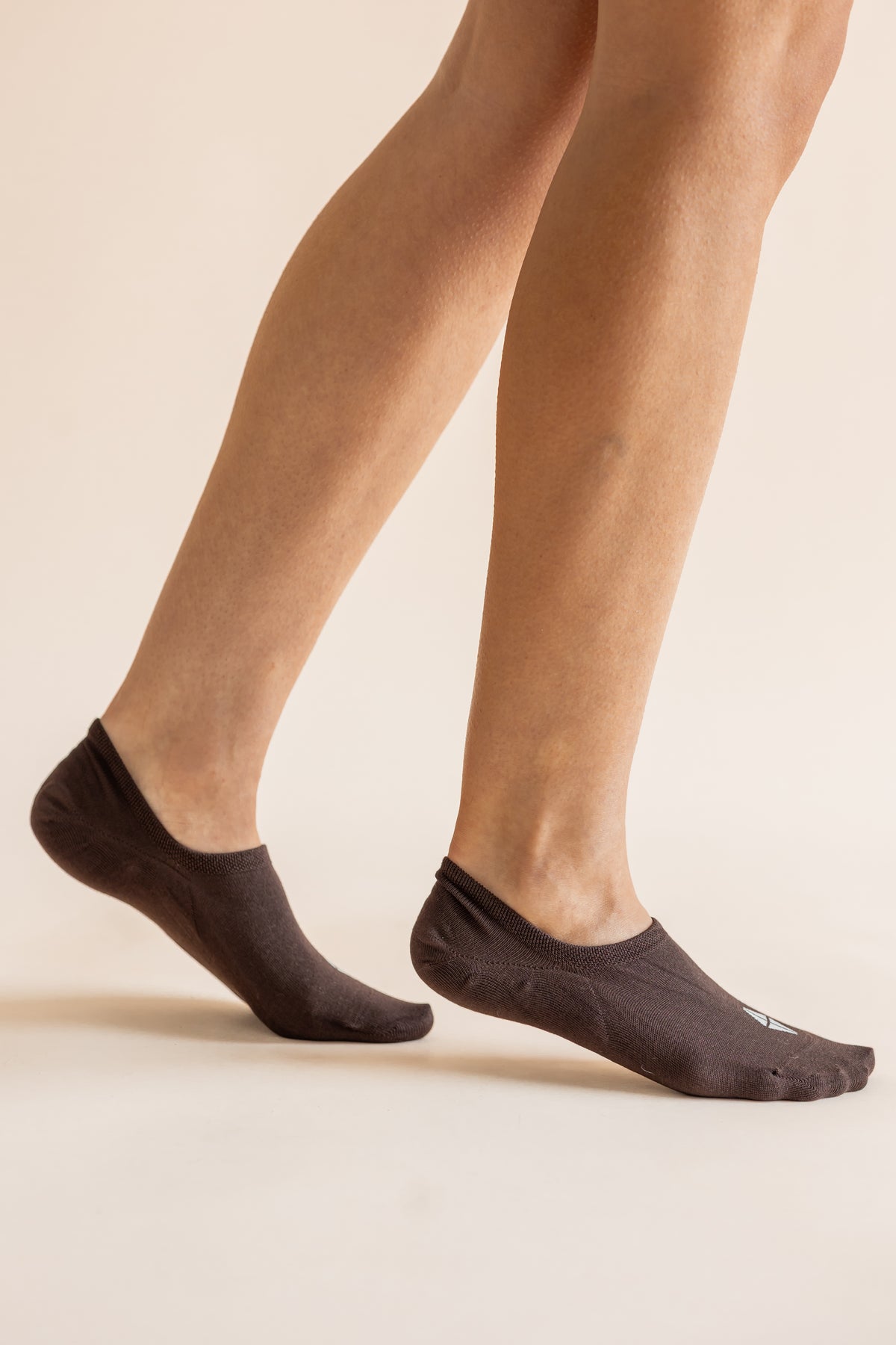 A close-up of a person's feet wearing dark brown low-cut no-show socks against a solid light beige background.