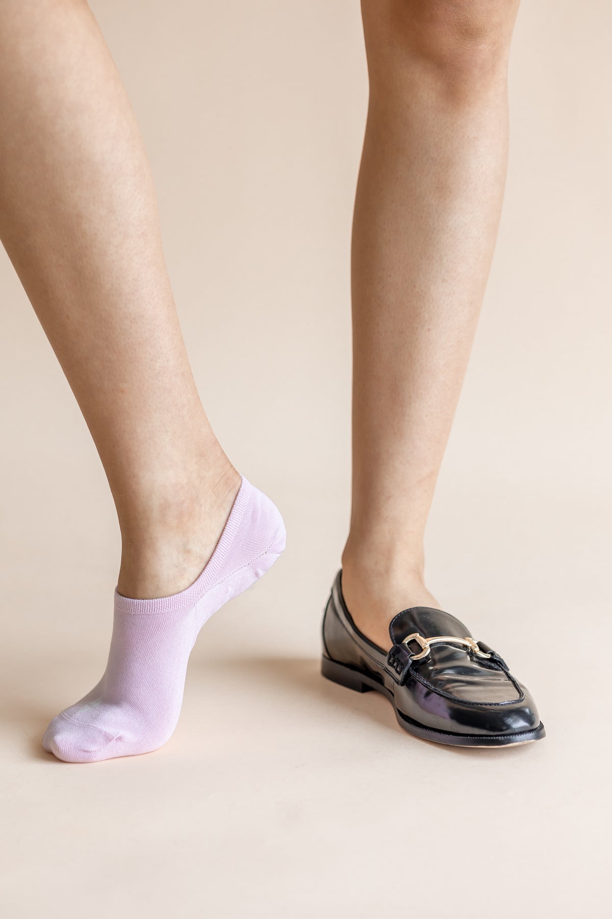 A person's legs shown from the calves down, wearing a light lavender no-show sock on one foot and a black patent leather loafer with a gold buckle on the other, against a solid beige background.