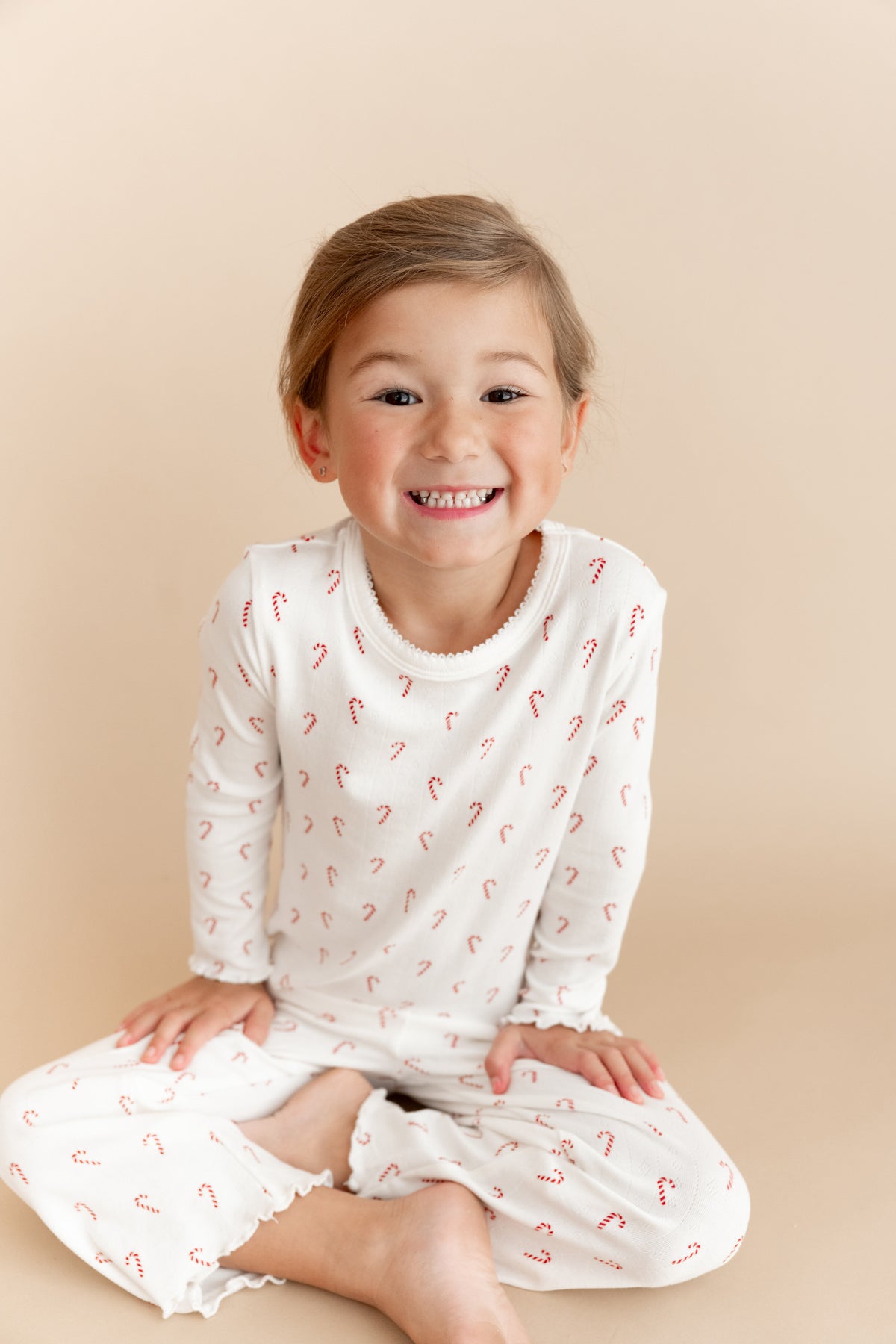 A young child smiling while wearing a white long-sleeve pointelle top and matching pants with a small red candy cane pattern, posed against a solid beige studio background.