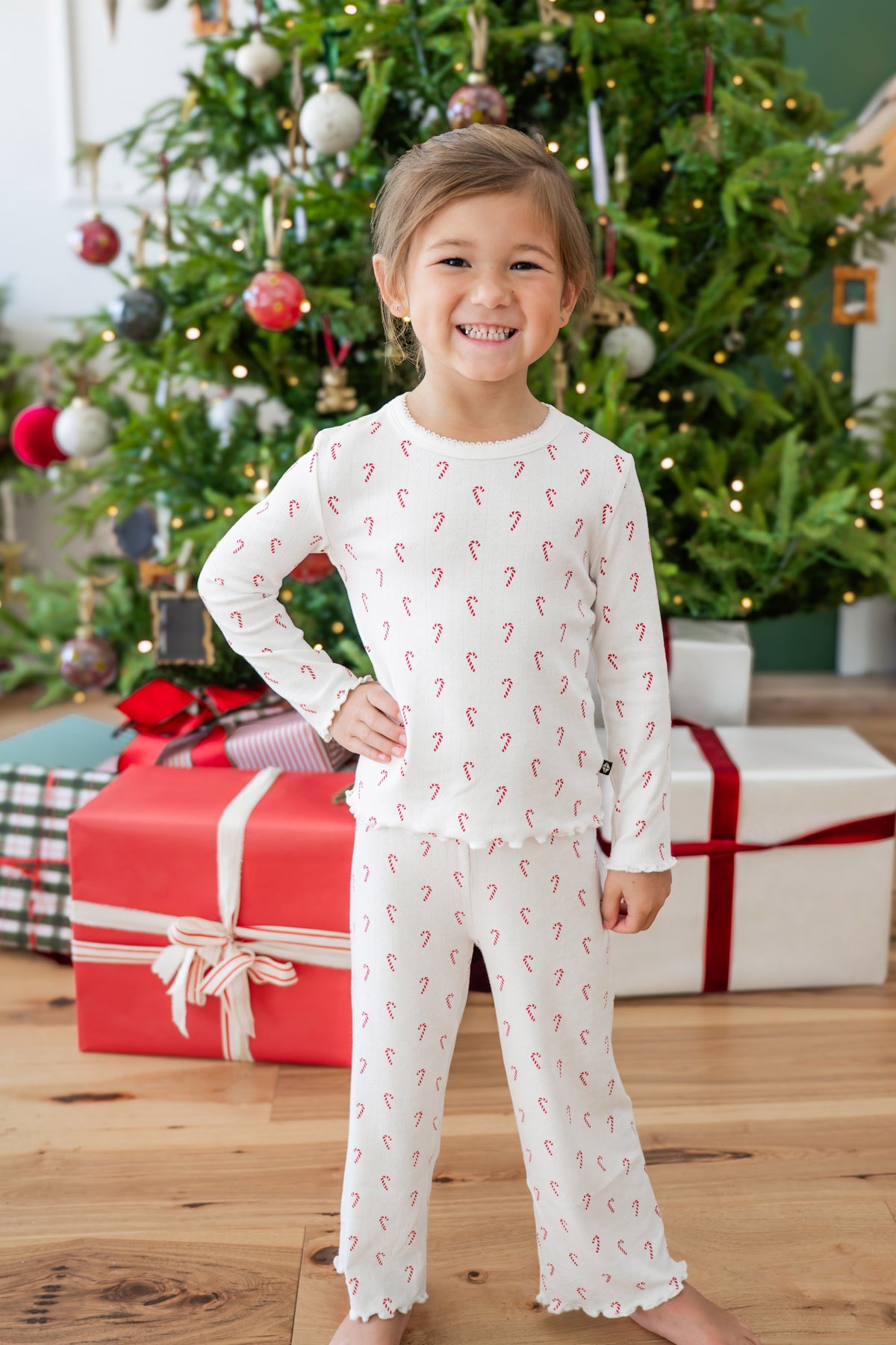 A young girl smiling in a festive holiday set featuring a white long-sleeve top and matching pants with a red candy cane print, posed in front of a decorated Christmas tree and wrapped gifts.
