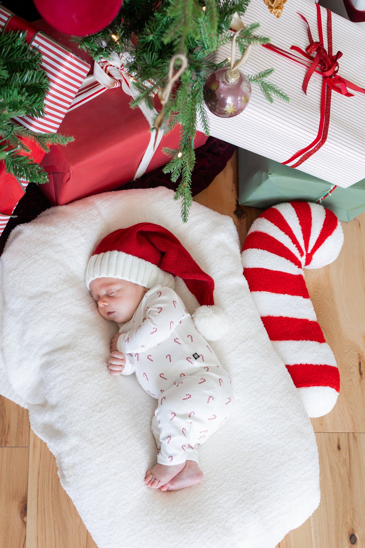 Baby sleeping wearing Pointelle Kimono Set in Candy Cane and red hat on a white sheet near a Christmas tree and presents