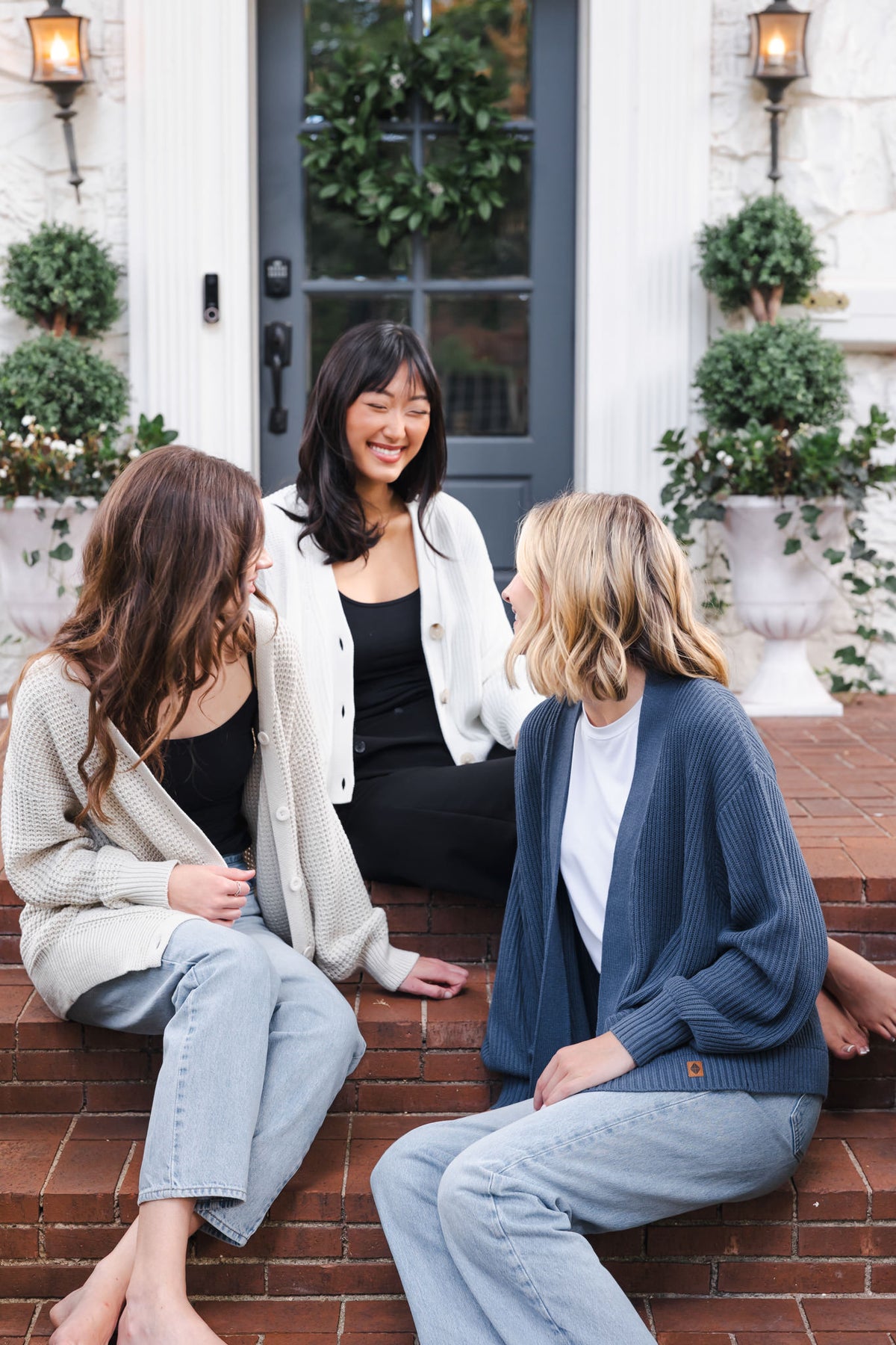 Three modal sitting outdoors wearing Chunky Knit Women's Cropped Cardigan in Cloud, Chunky knit women's waffle cardigan in almond and chunky knit oversized cardigan in oxford