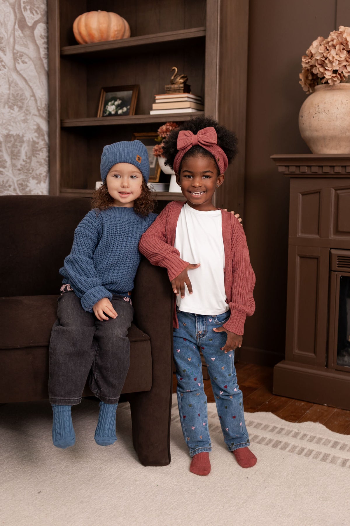 Two girls in a living room wearing Chunky Knit Sweater in Oxford with chunky knit pom beanie in oxford and chunky knit oversized cardigan in ember with chunky knit bow headband in ember