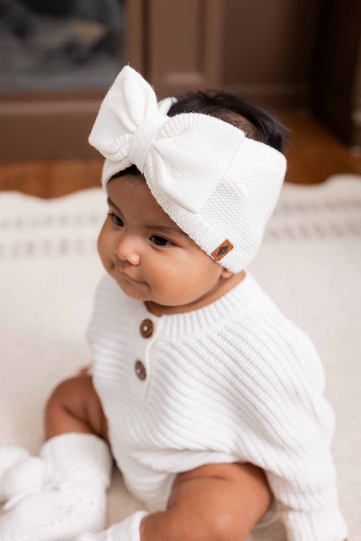 Baby wearing Chunky knit romper, Chunky knit bow headband and knit booties in cloud while sitting on a white carpet