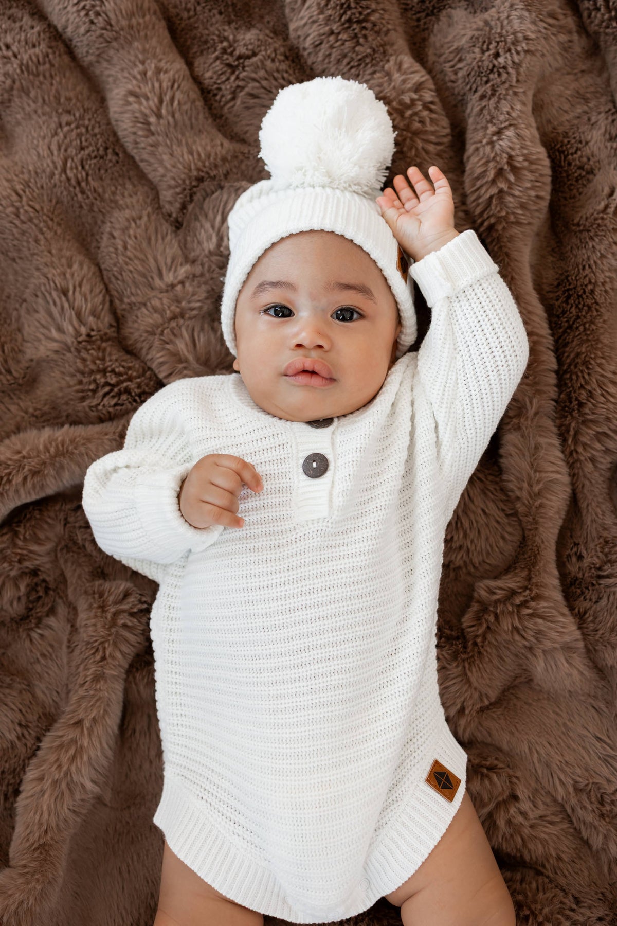 Baby wearing Chunky Knit Romper and chunky knit pom beanie in cloud lying on brown fur blanket