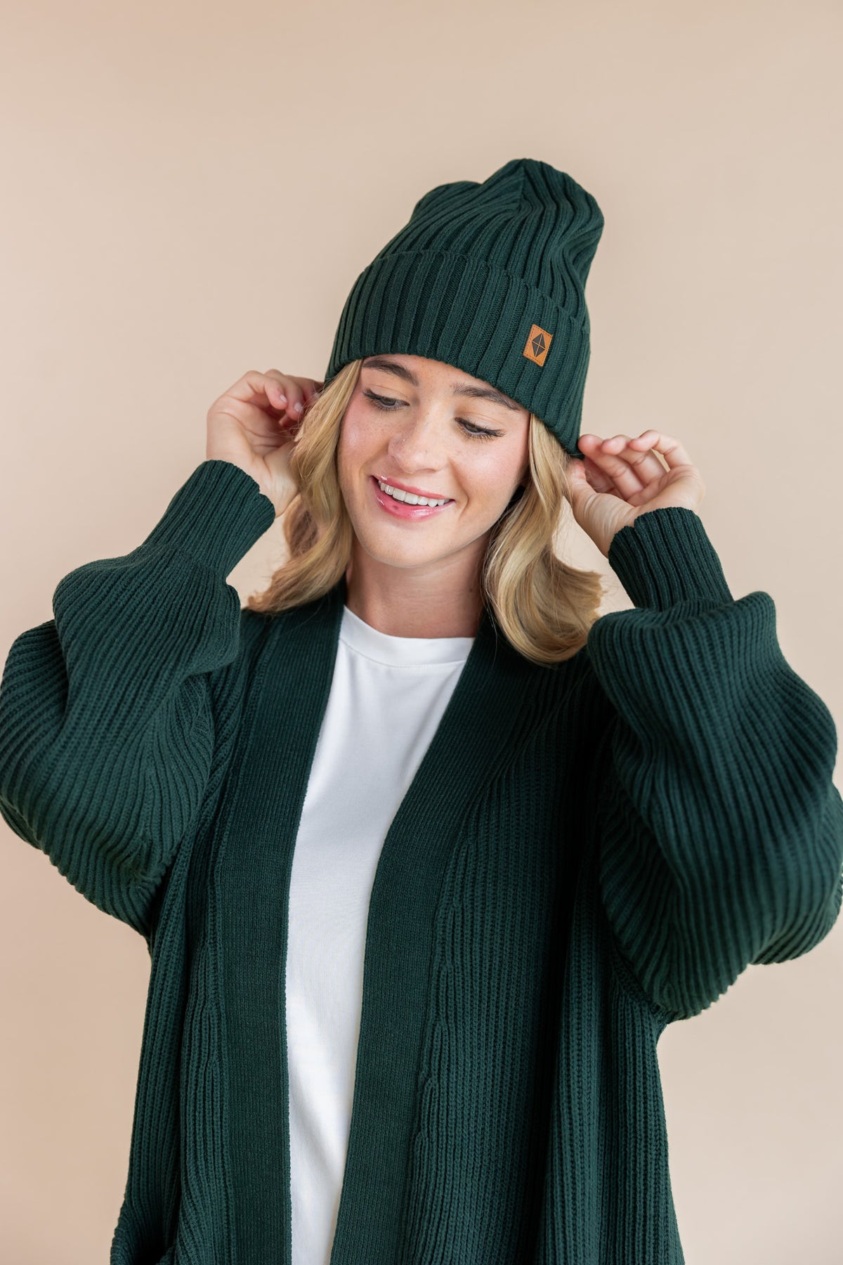 Modal posing in chunky knit women's oversized cardigan and chunky knit adult ribbed beanie in cypress on a beige background