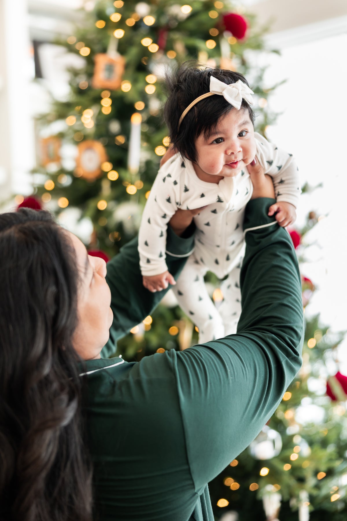 Mommy wearing long sleeve pajama set in cypress holding her Baby wearing Pointelle Zippered Footie in Christmas Trees with a Christmas tree in the background