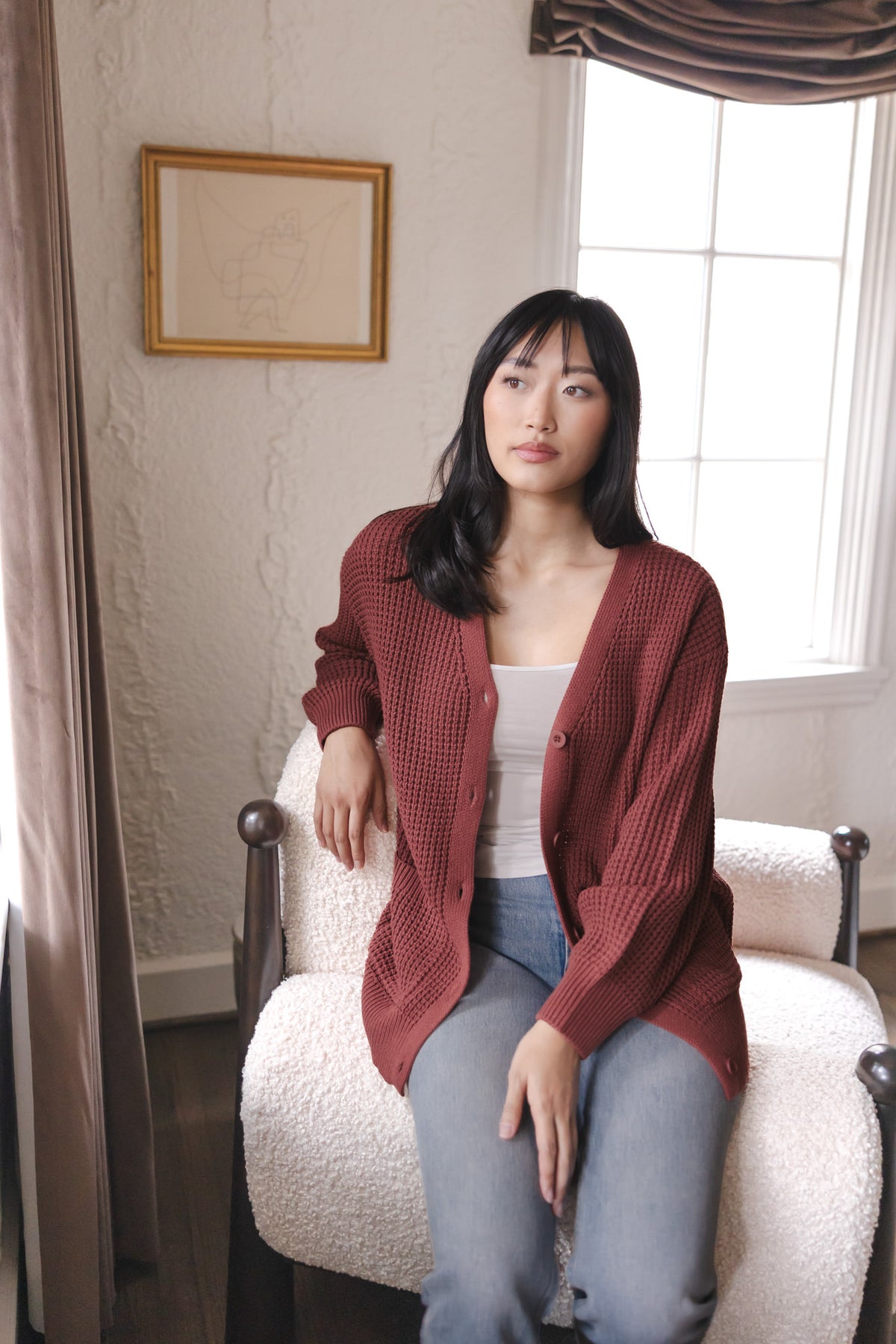 Modal sitting on a white sofa wearing Chunky Knit Women's Waffle Cardigan in Ember over white camisole and blue jeans