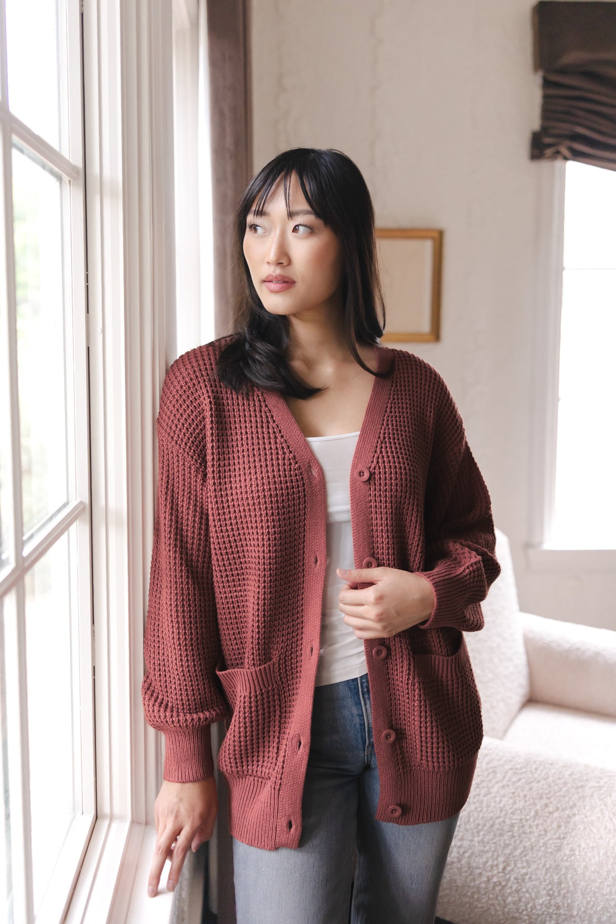 Modal standing against a window wearing Chunky Knit Women's Waffle Cardigan in Ember over white camisole and blue jeans