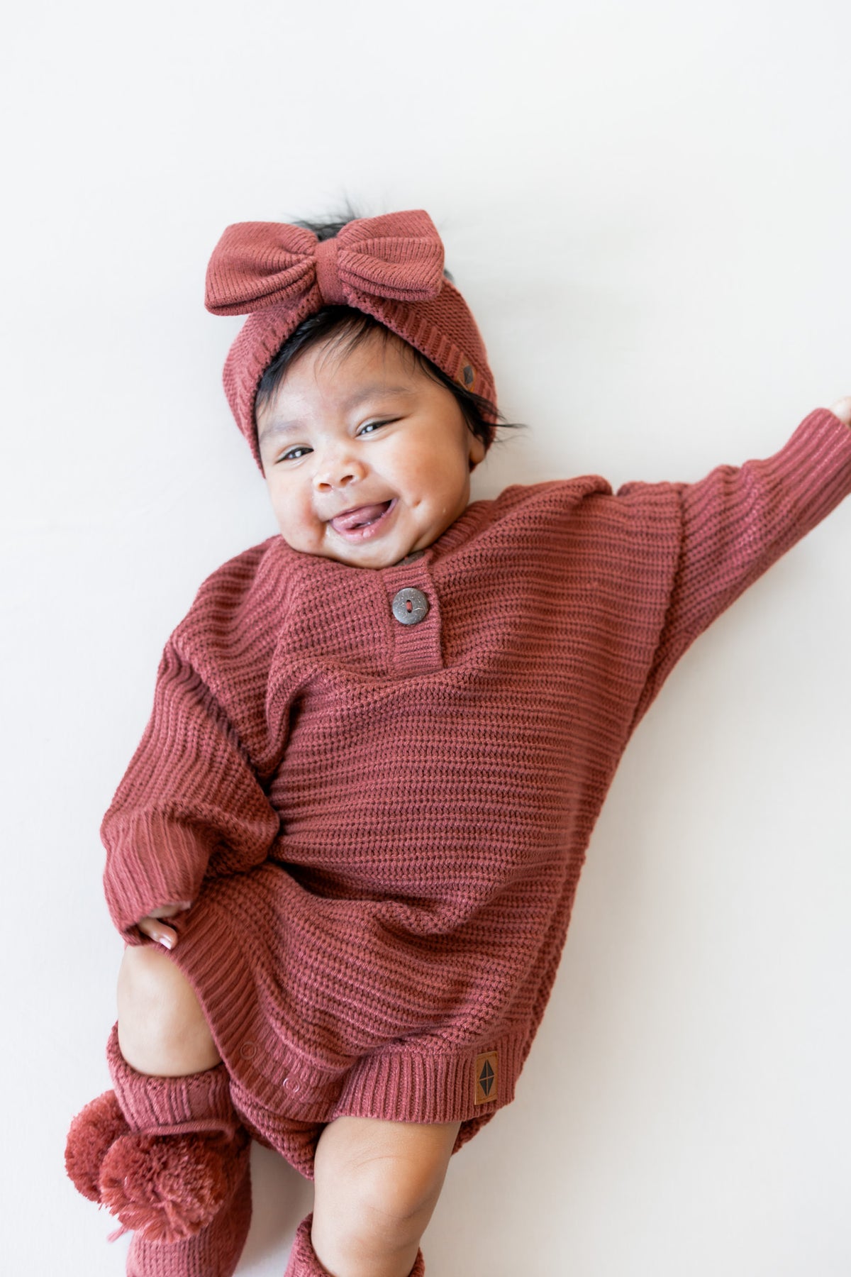 Baby wearing Chunky Knit Romper, chunky knit bow headband and knit booties in Ember on a white background