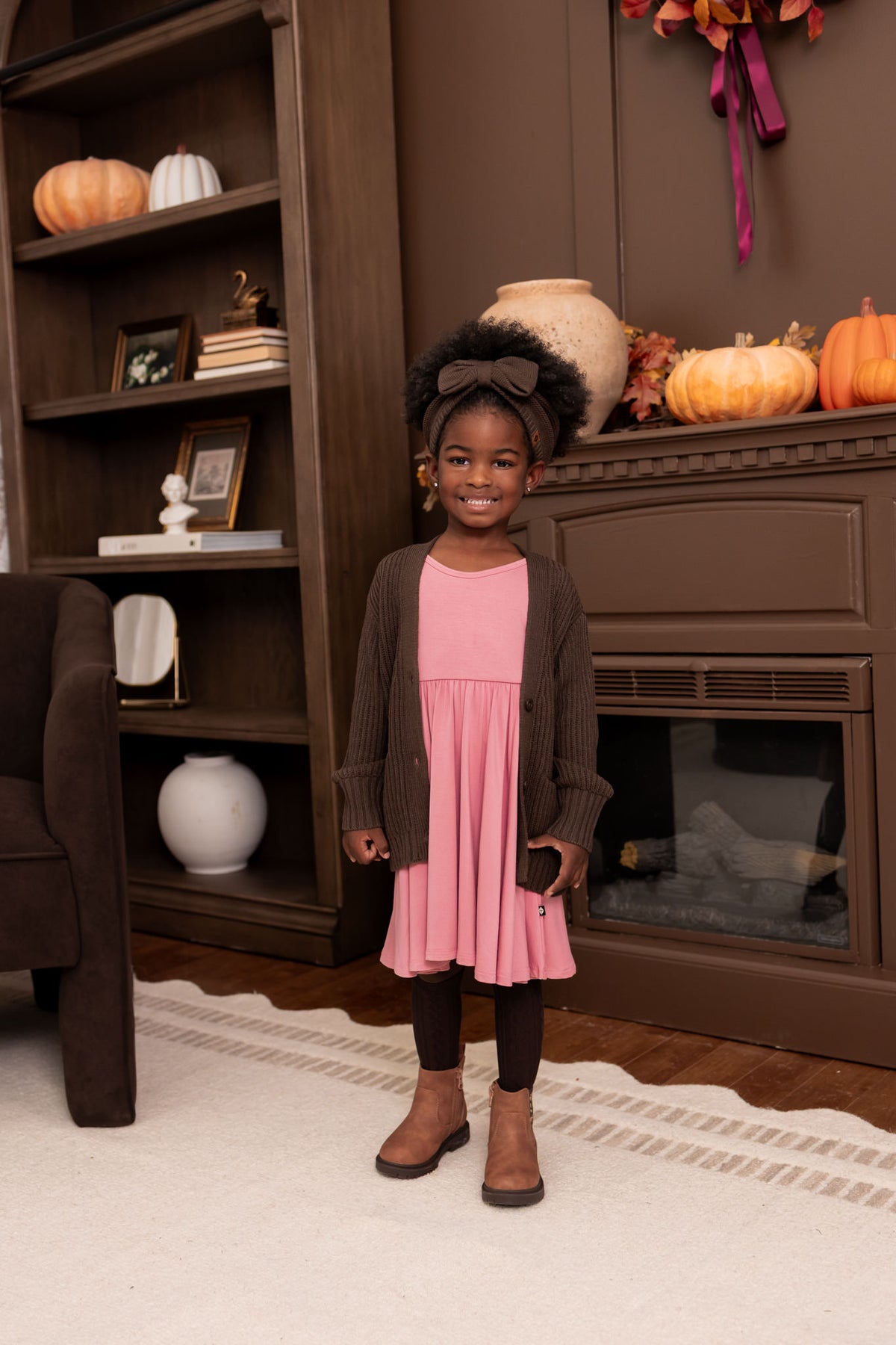 Girl wearing Chunky Knit Oversized Cardigan in Espresso over Twirl dress in pink and chunky knit bow headband in espresso standing in a living room
