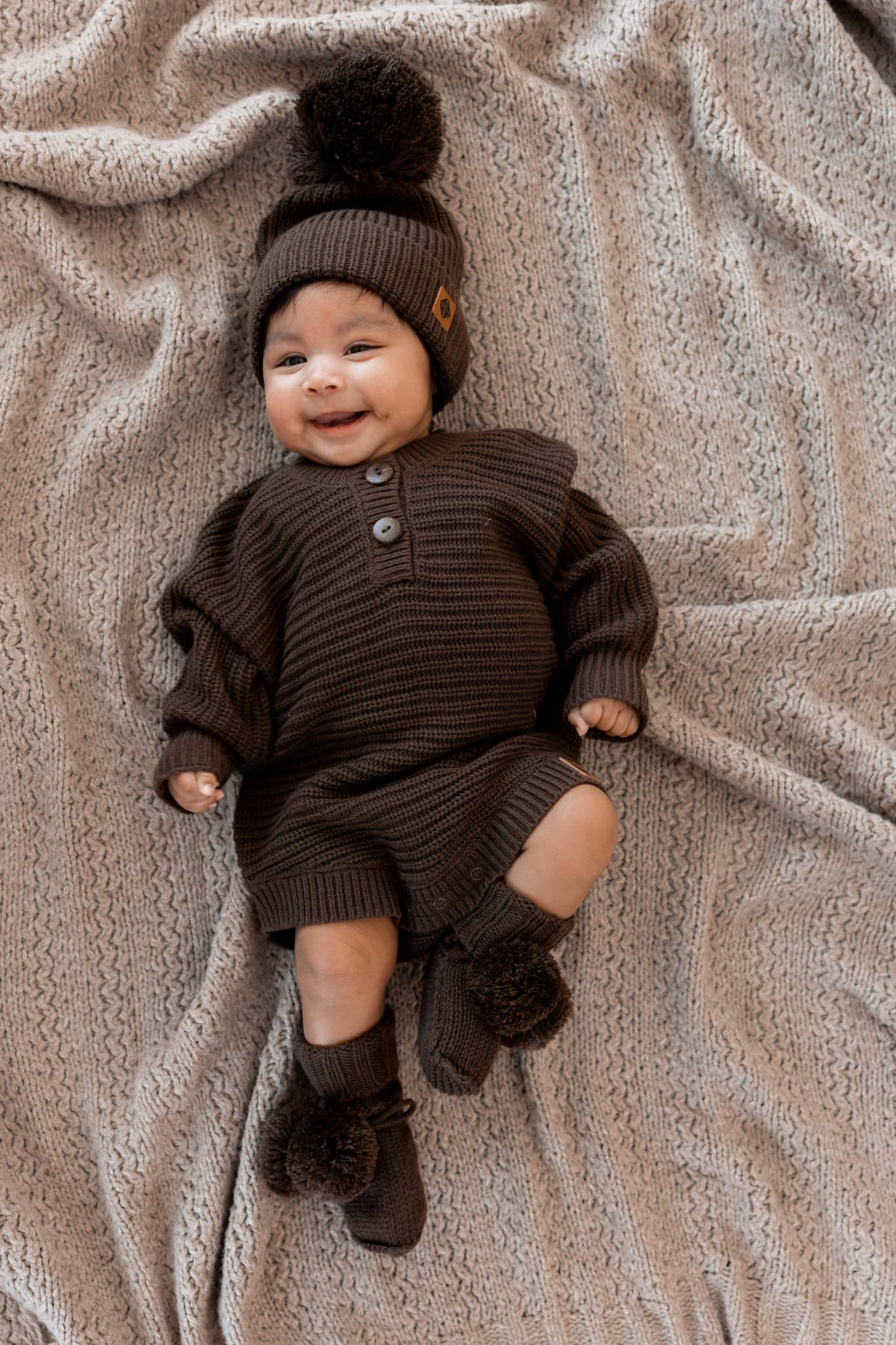 Baby wearing Chunky Knit Romper, chunky knit pom beanie and knit booties in Espresso on a beige blanket