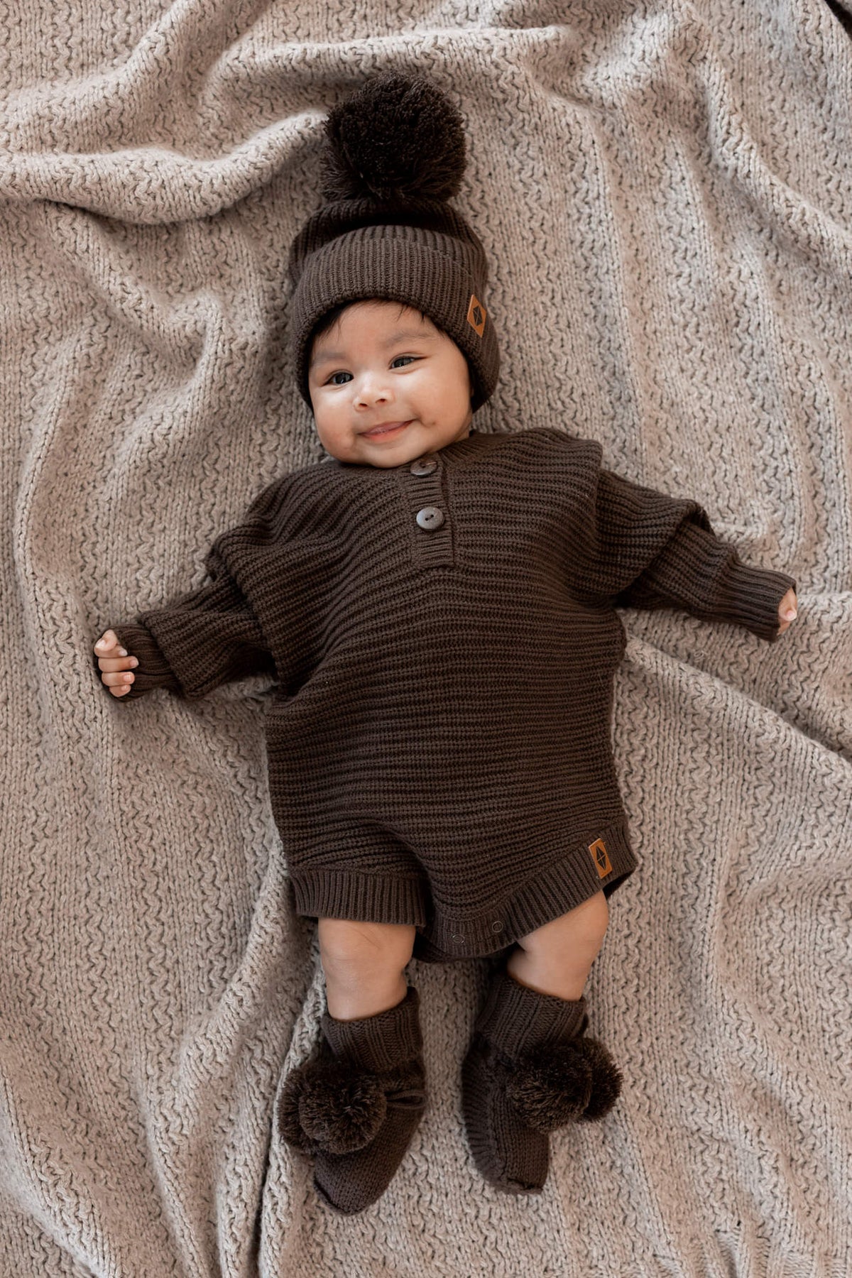 Baby smiling in Chunky Knit Romper, chunky knit pom beanie and knit booties in Espresso on a beige blanket