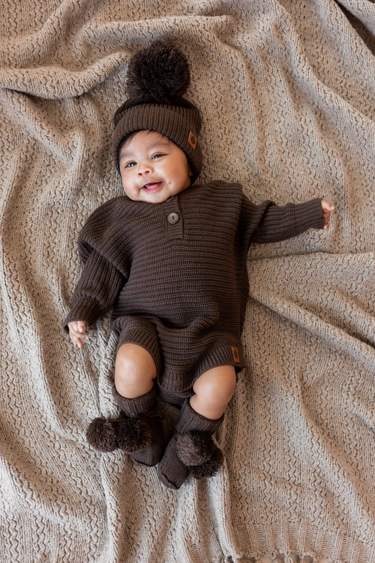 Baby wearing Knit Booties, chunky knit romper and chunky knit pom beanie in Espresso lying on a beige blanket