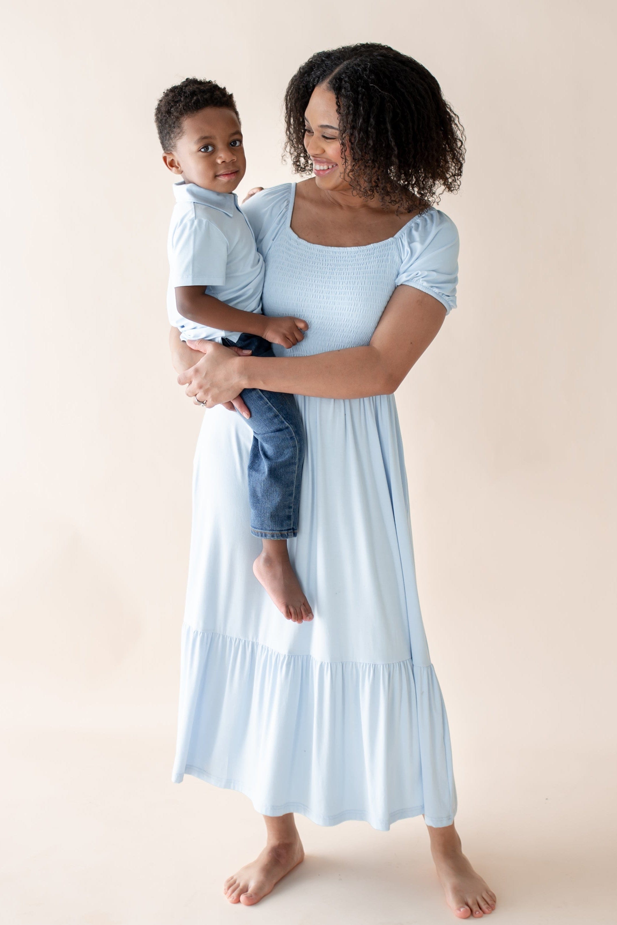 Woman in a light blue dress holding a child wearing matching blue polo against a beige background
