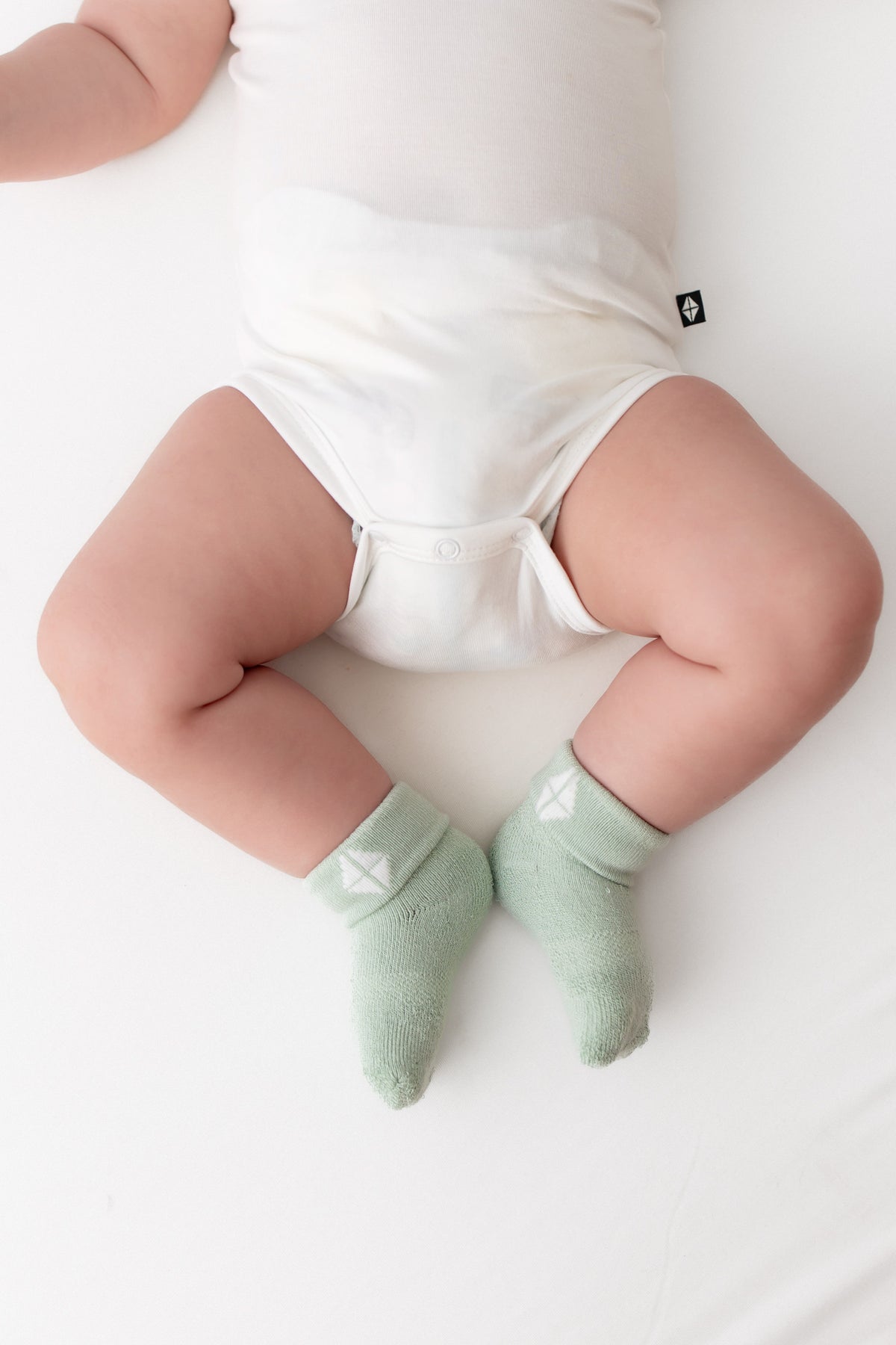 A baby lying on a white surface wearing pale mint green socks with a small white geometric logo, along with a white bodysuit.