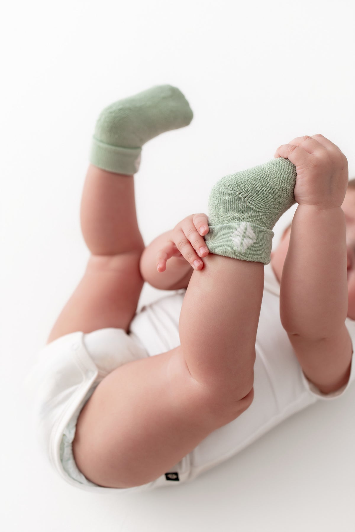 A baby lying on their back wearing light sage green knit socks with white trim, paired with a white bodysuit, against a clean white background.
