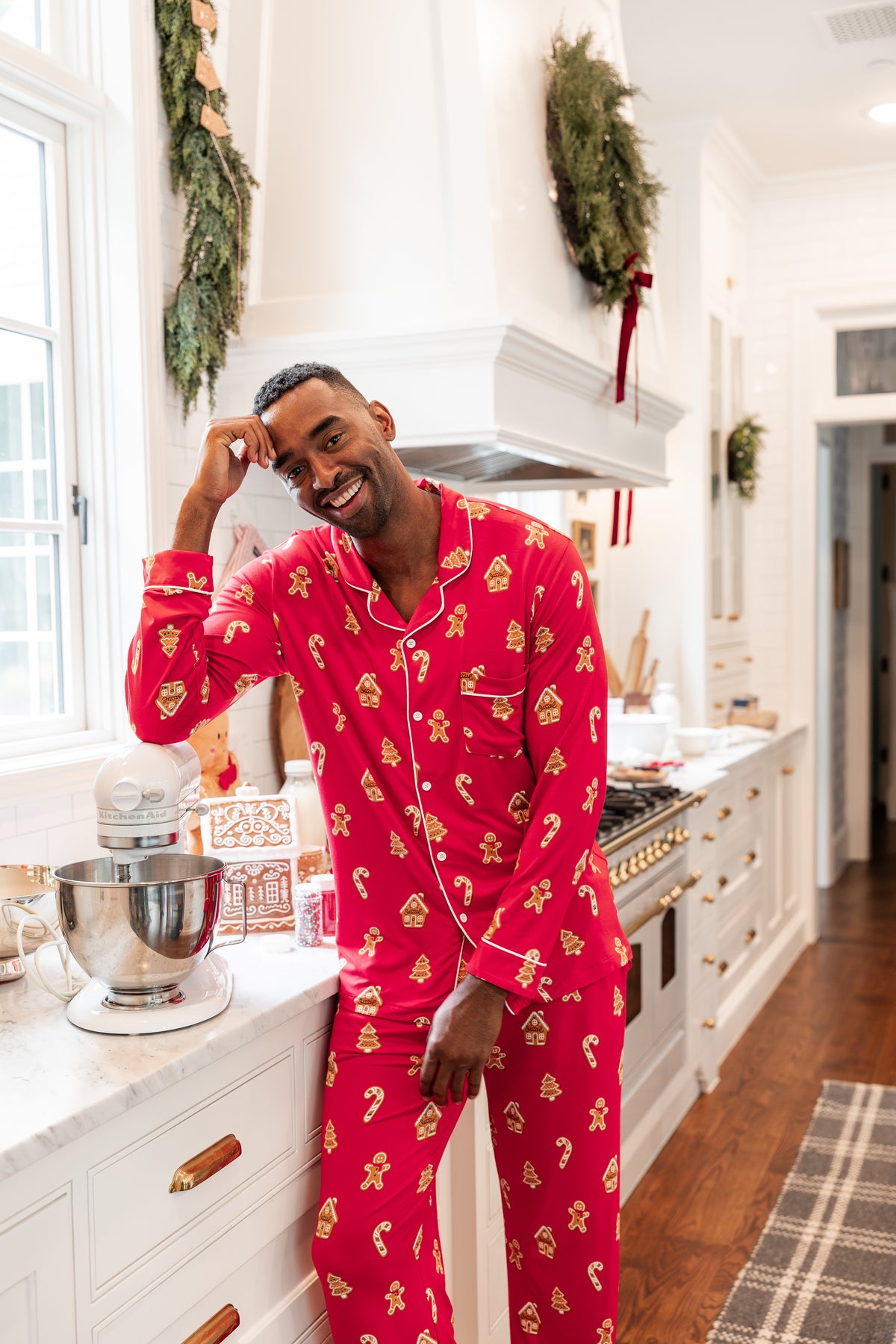 A man wearing a festive red and gold Christmas-themed pajama set stands in a bright white kitchen, smiling and posing next to a countertop with a stand mixer.