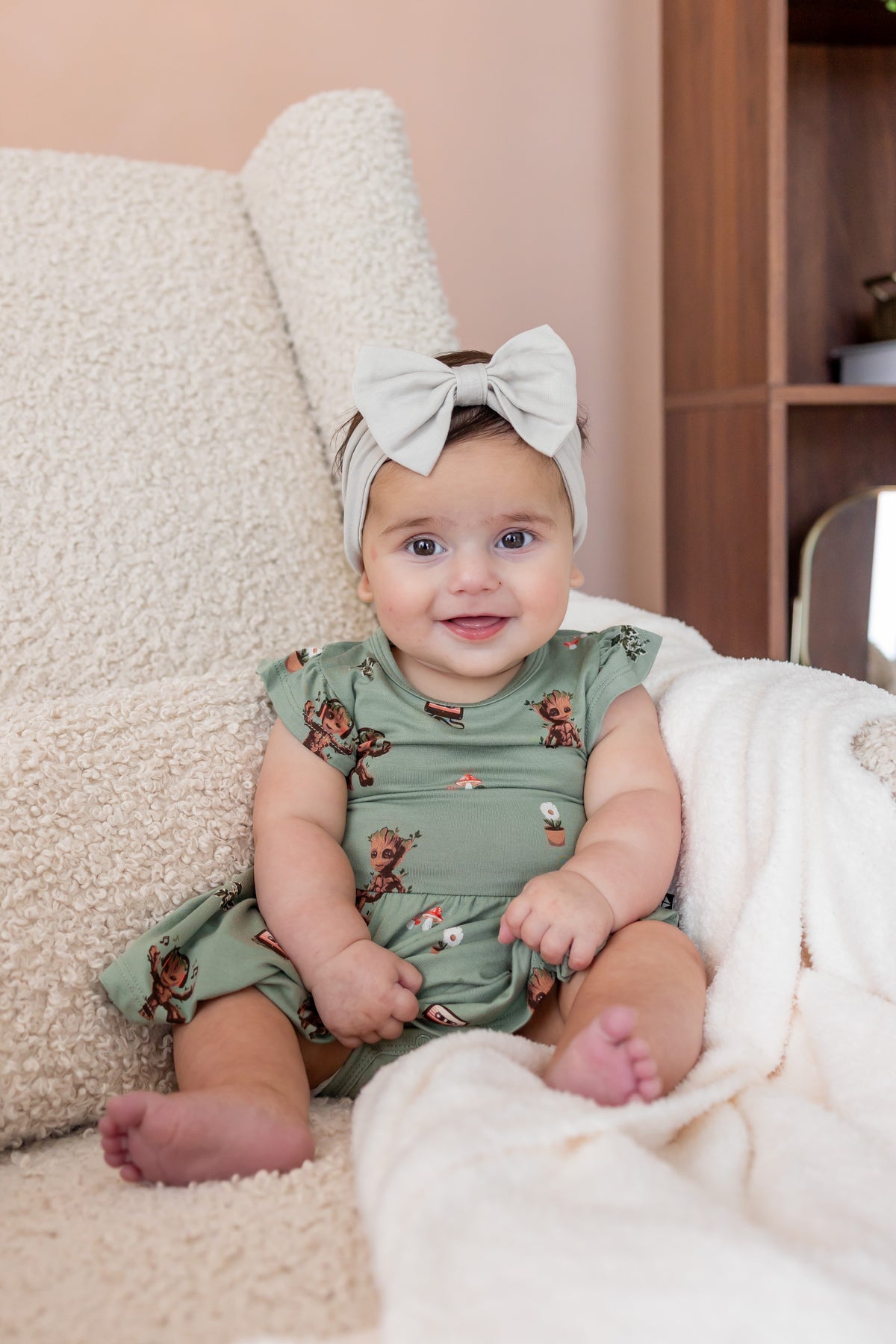 Baby sitting upright on a white sofa wearing Green Twirl Bodysuit Dress in Groot print and a beige knotted bow