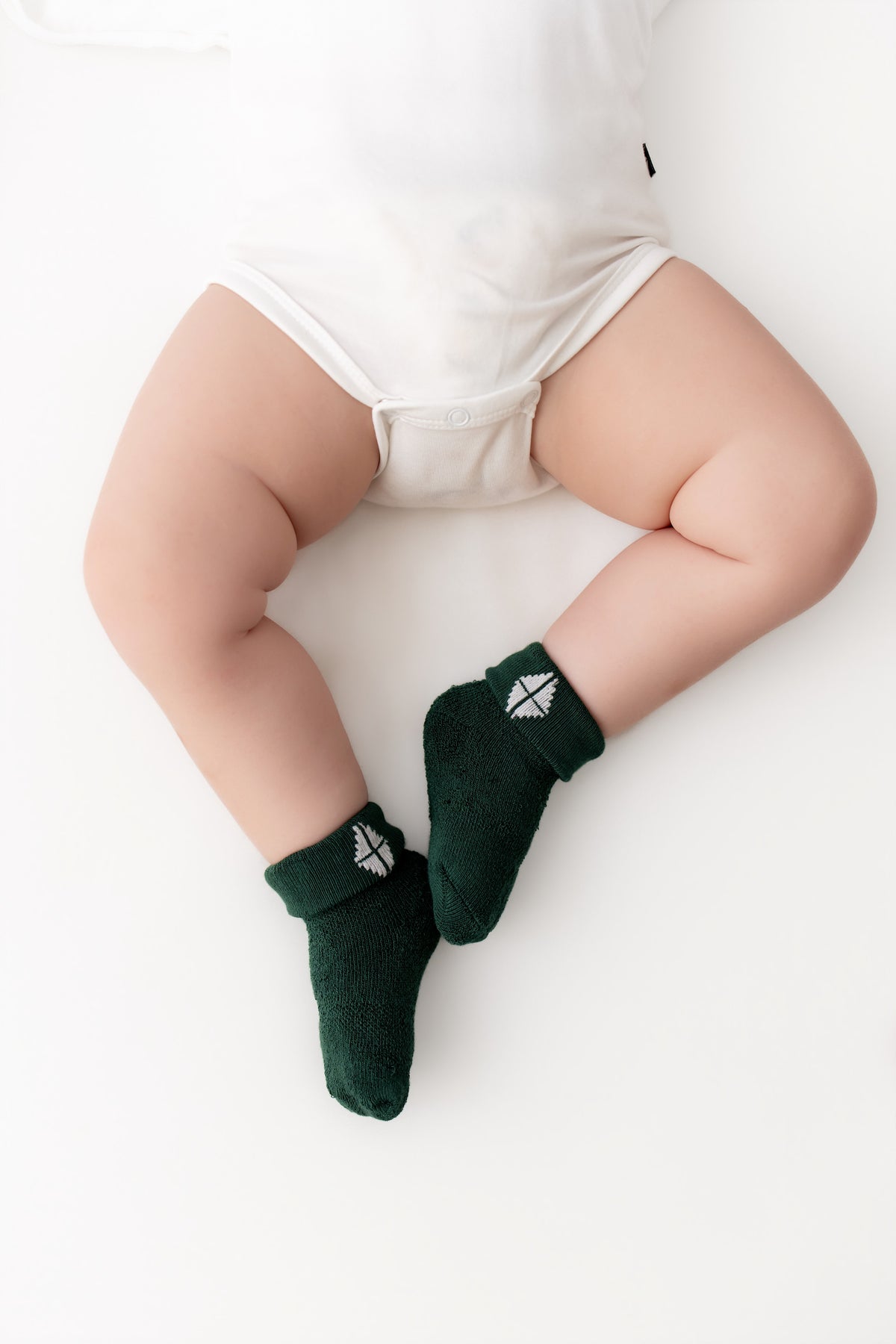 A baby's legs wearing dark green knit socks with a small white geometric logo, styled with a white bodysuit on a clean white background.