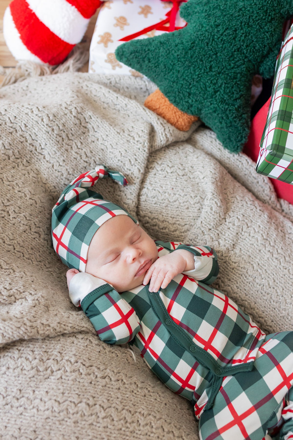 A sleeping newborn baby wearing a green, red, and white plaid kimono set and matching hat, lying on a cozy beige knit blanket with Christmas-themed toys in the background.