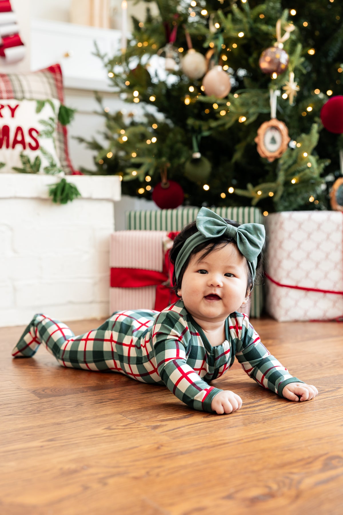 A baby wearing a green, red, and black plaid footed onesie and a matching green bow headband lies on a wooden floor in front of a decorated Christmas tree and wrapped gifts.