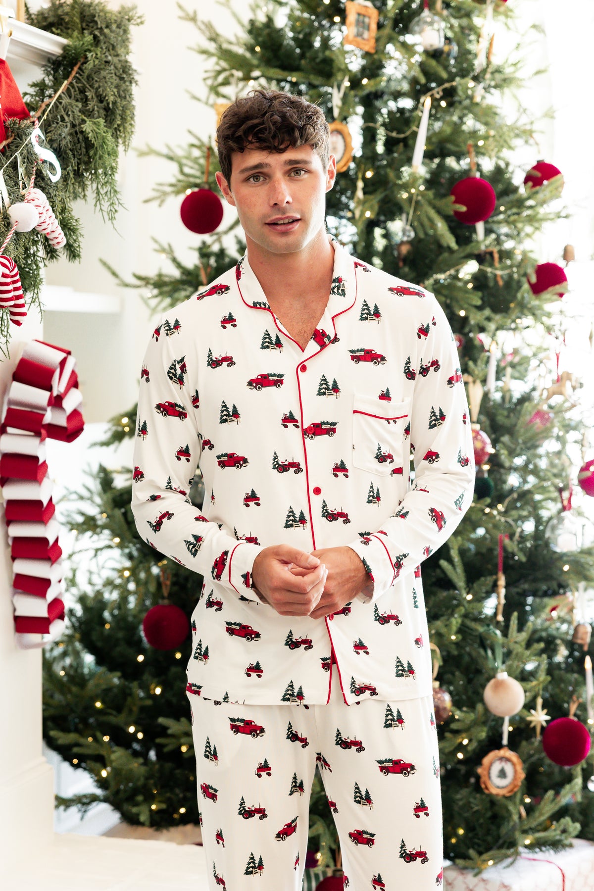 A man wearing a white long-sleeve pajama set with a red and green Christmas truck and tree print, standing in front of a decorated Christmas tree.