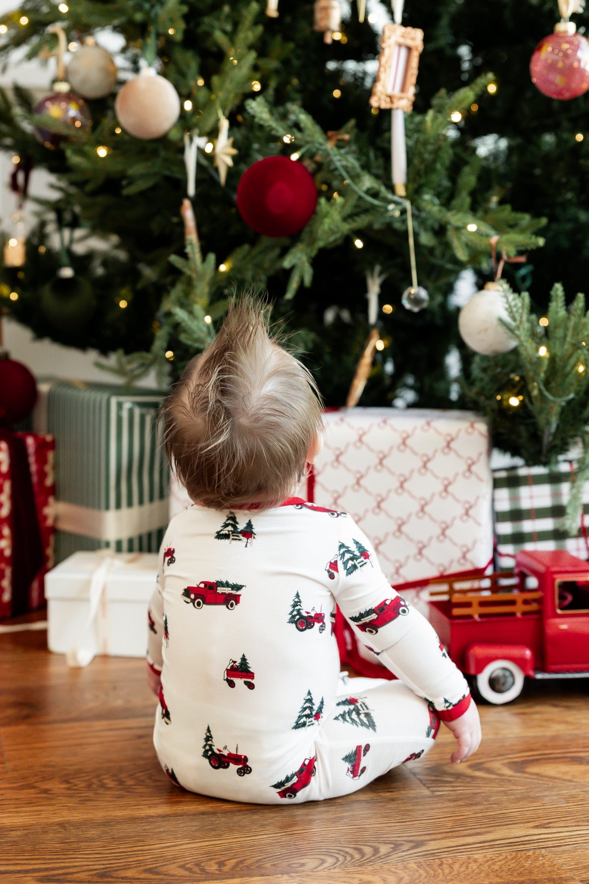 A toddler sits on a wooden floor in front of a decorated Christmas tree, wearing a white long-sleeve pajama top with a red and green truck and tree print. Gifts and a red toy truck are visible around the tree.