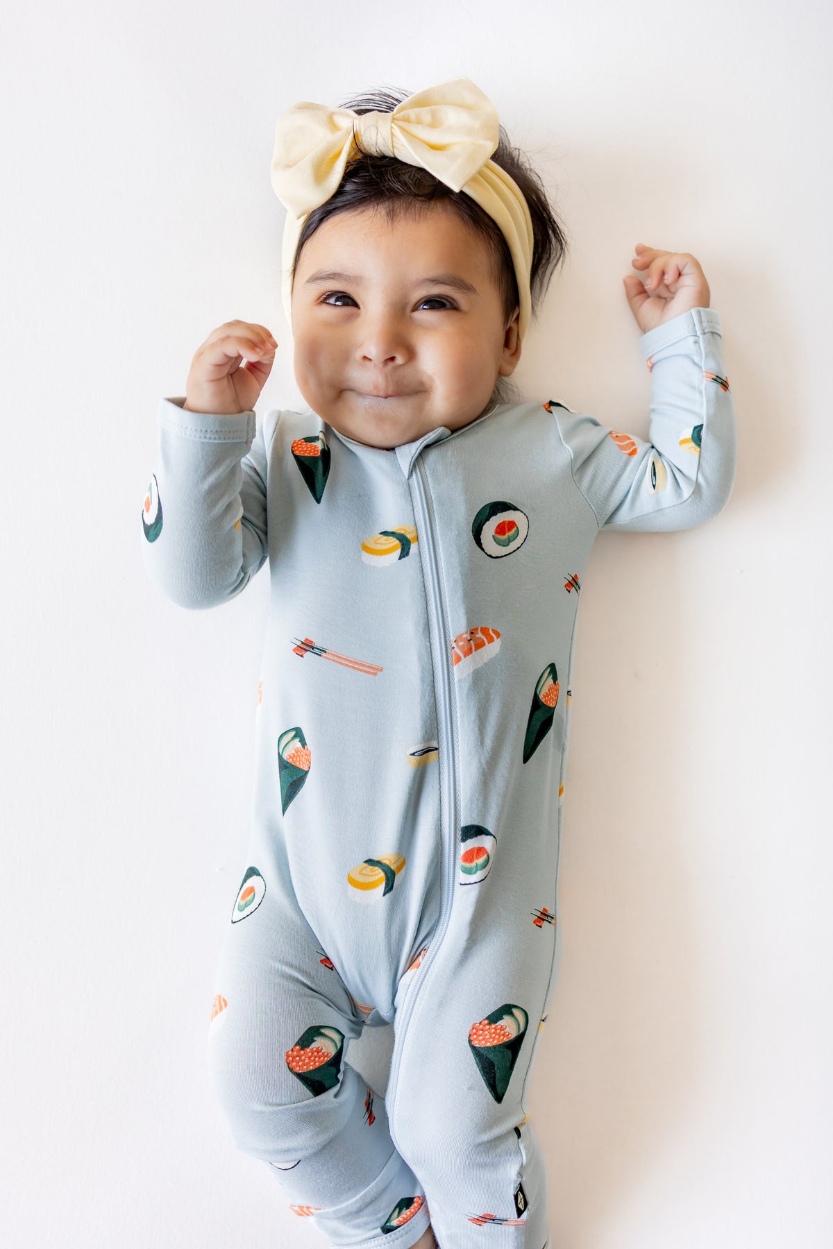 Baby laying down with a white background wearing ice sushi pajama set and a yellow bow