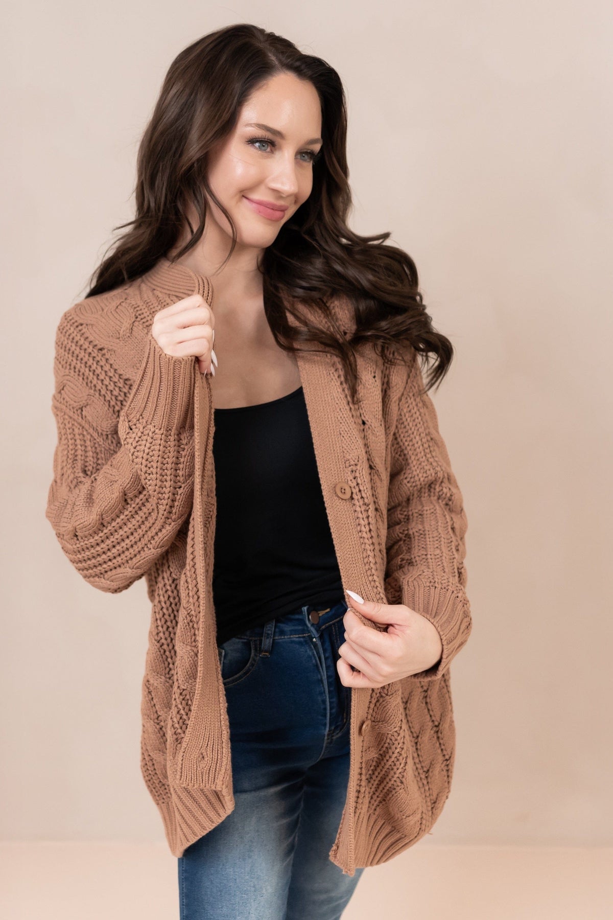 Female model standing wearing the Cable Knit Adult Cardigan in Latte holding the openings of the cardigan in both of her hands