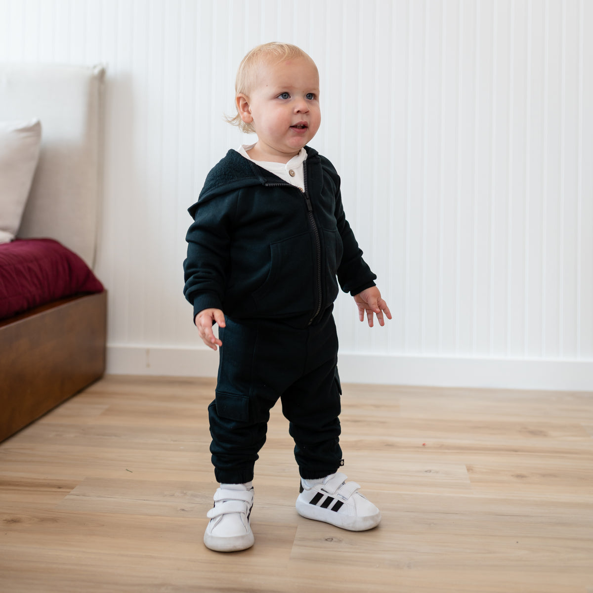 Young toddler standing beside a bed wearing the Fleece Zip Up Hoodie in Midnight and matching fleece cargo pant