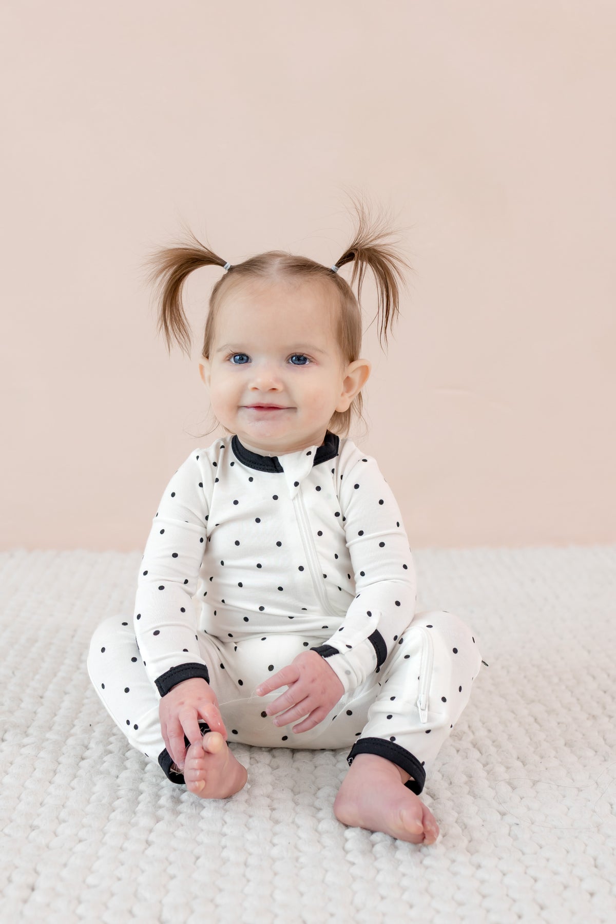 Child sitting in zippered romper with midnight polka dot print on beige background.