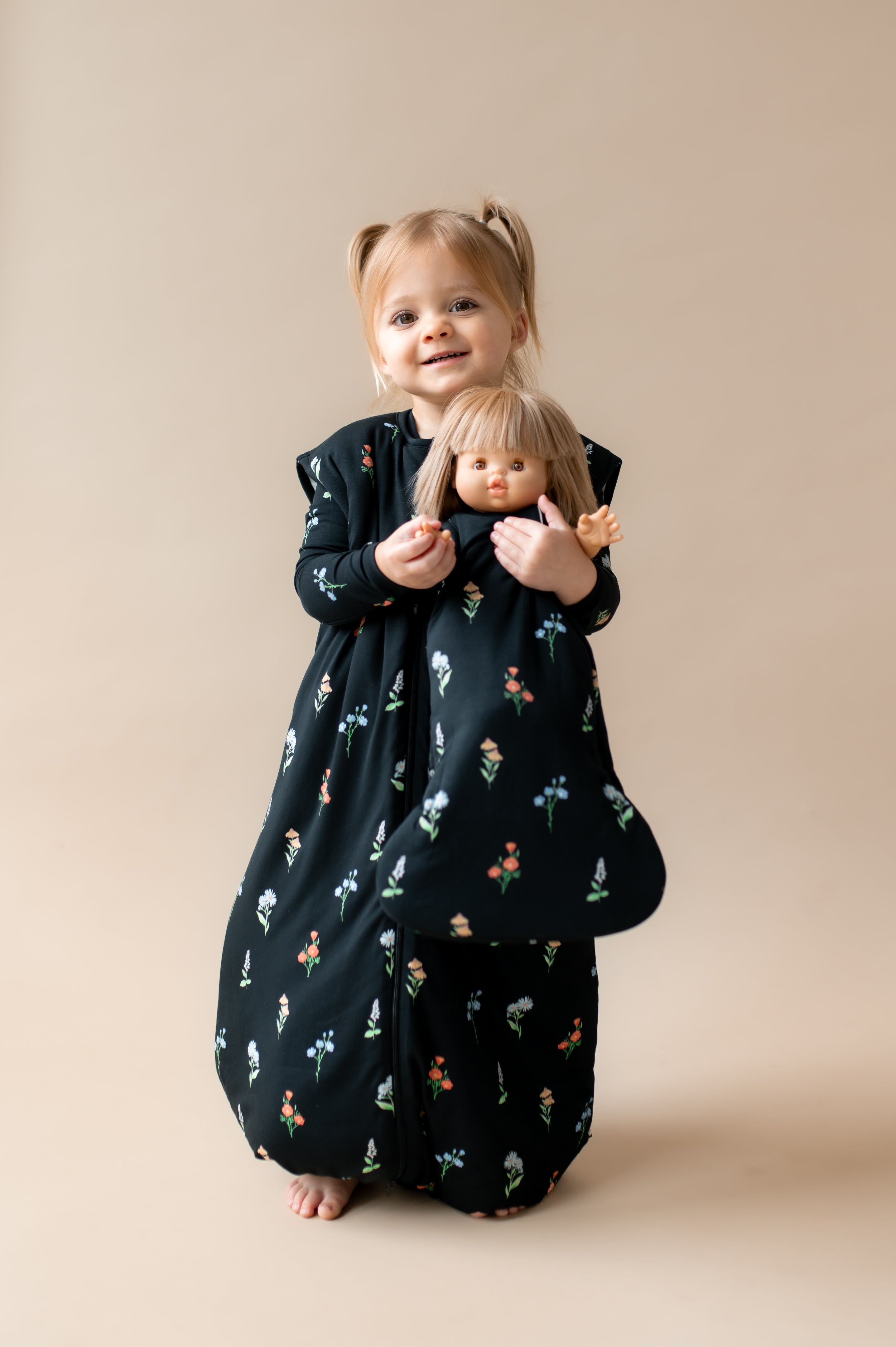Girl posing in Sleep Bag Walker in Midnight Wildflower 2.5 print holding a similar print baby doll sleep bag on a beige background