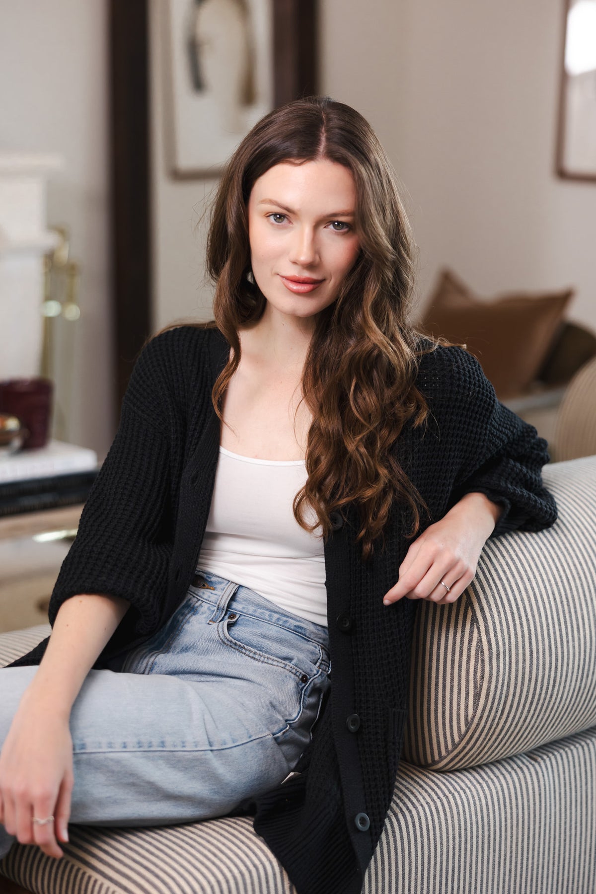 Modal sitting on a sofa wearing Chunky Knit Women's Waffle Cardigan in Midnight over white camisole and blue jeans