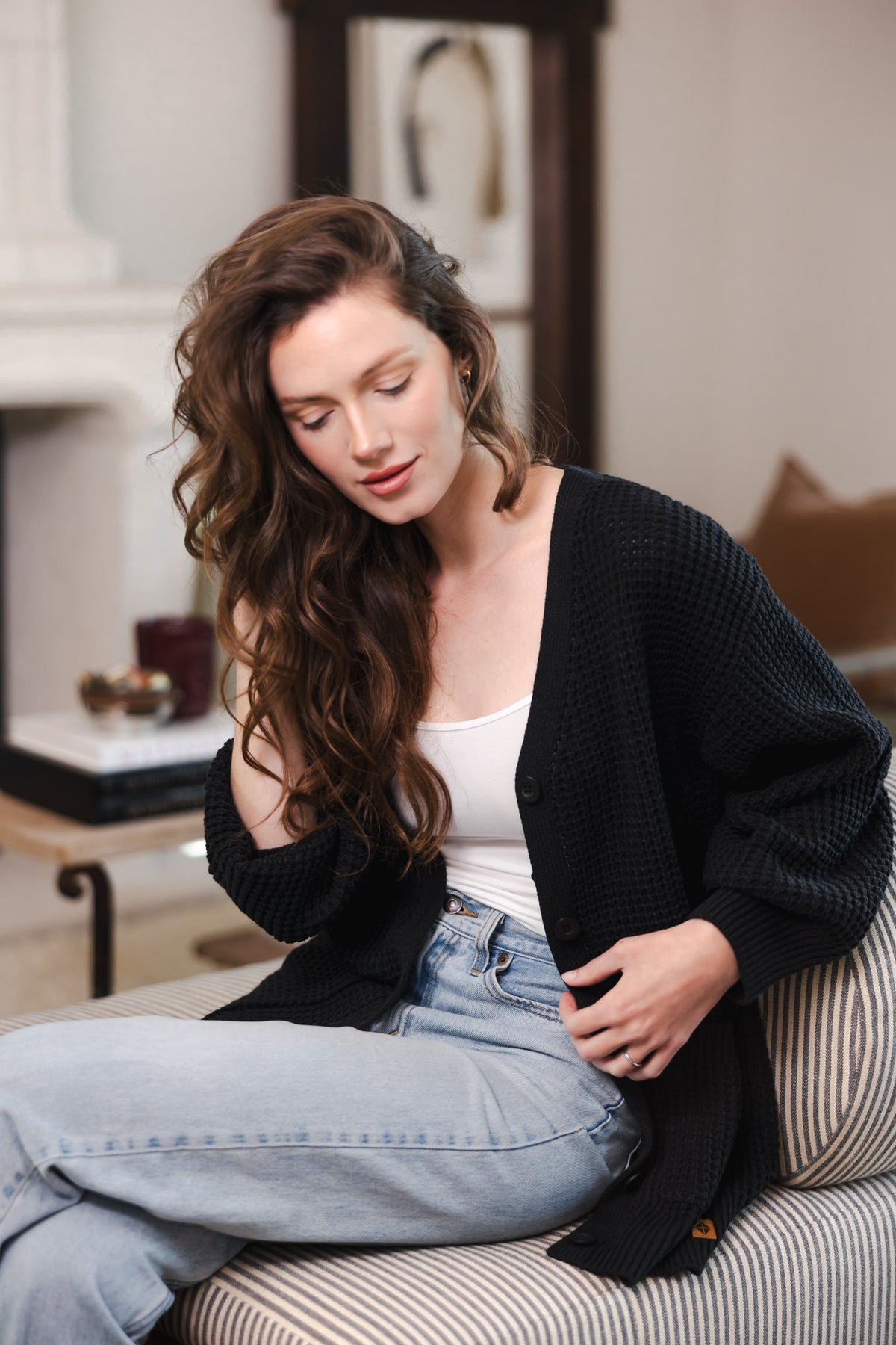 Modal sitting on a sofa posing in Chunky Knit Women's Waffle Cardigan in Midnight over white camisole and blue jeans