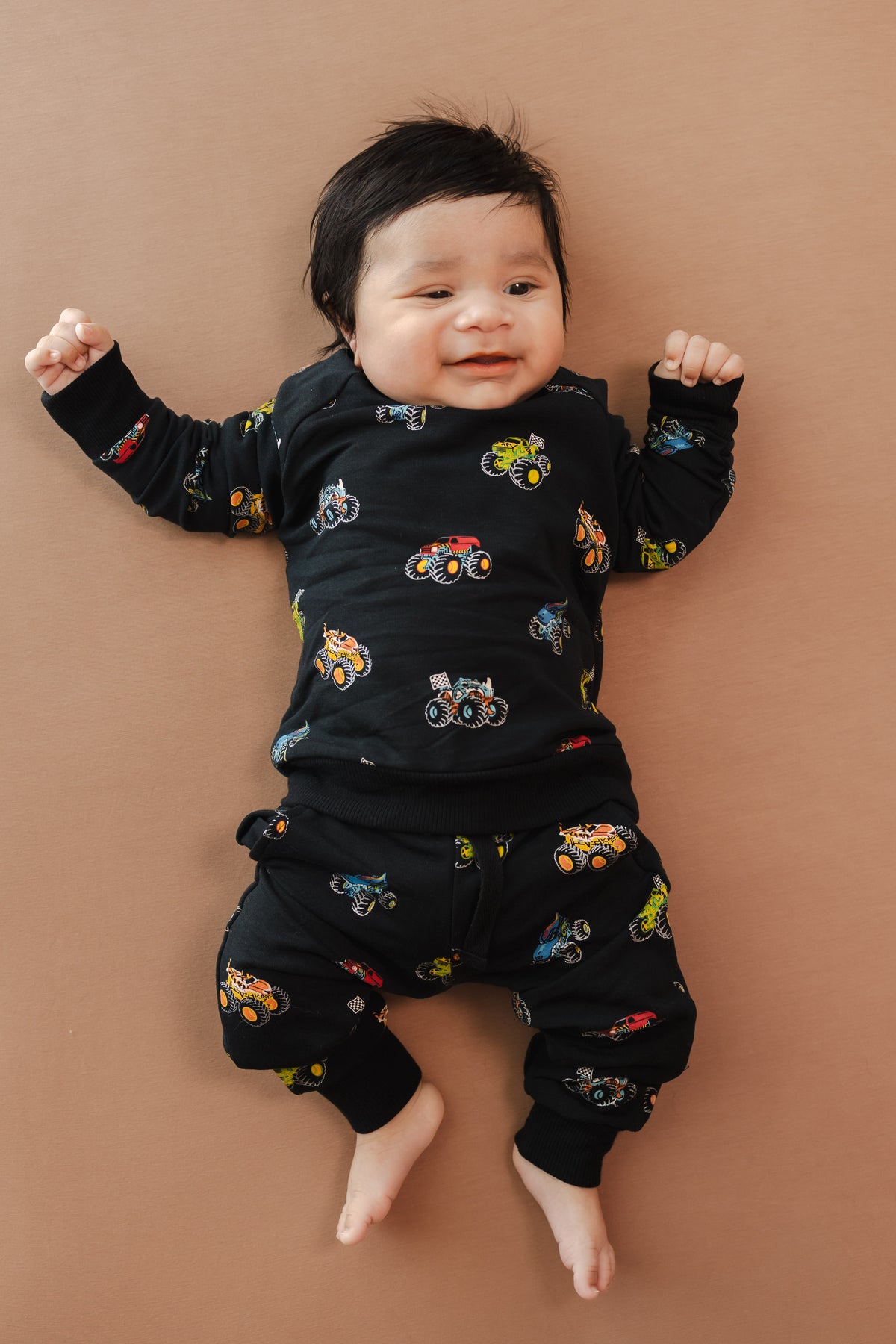 Baby wearing black Long Sleeve Jogger Set in Monster Truck print on beige background