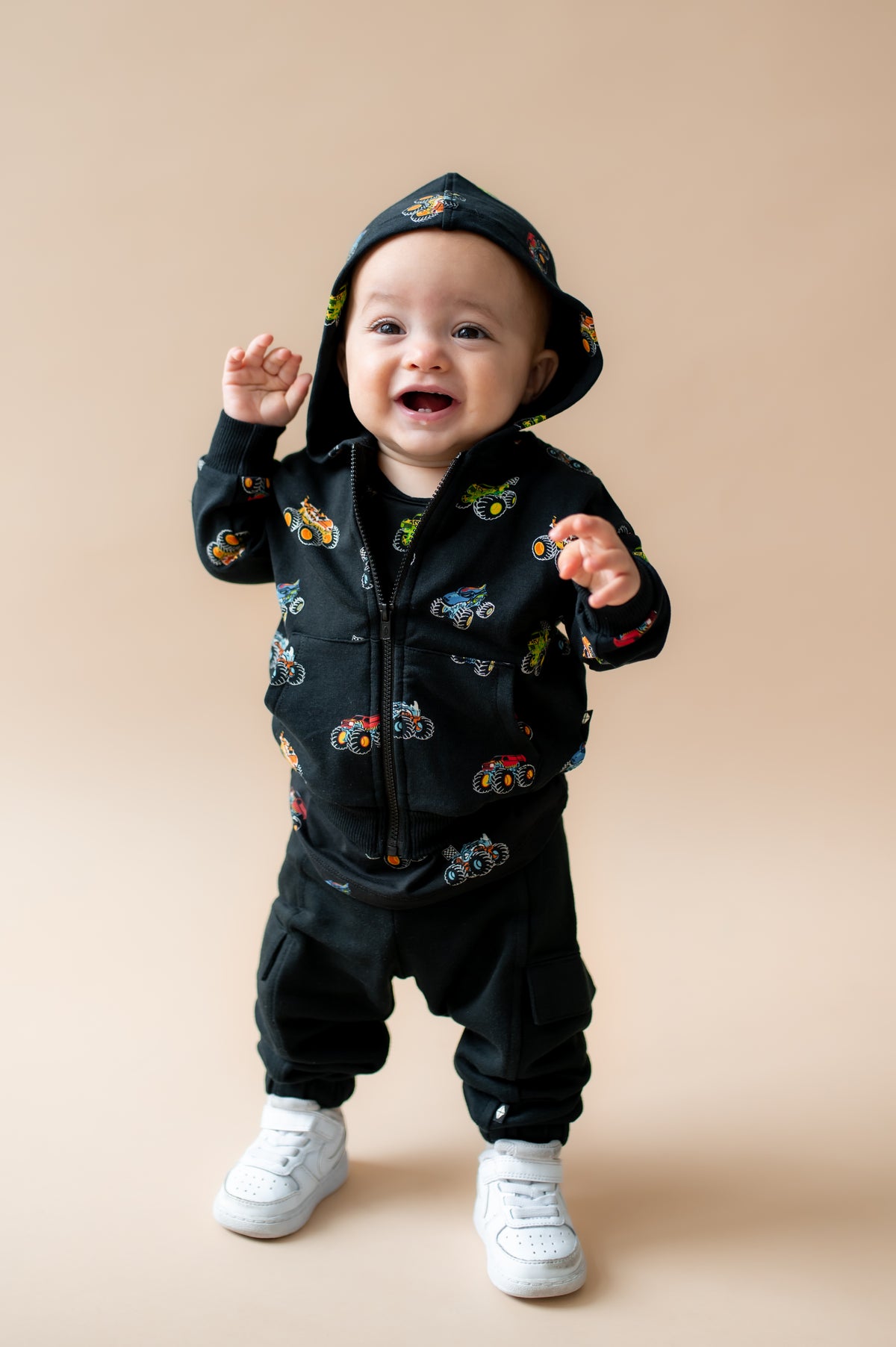 Toddler wearing Black Zip-Up Hoodie in Monster Truck print on black pant and white shoes on a beige background