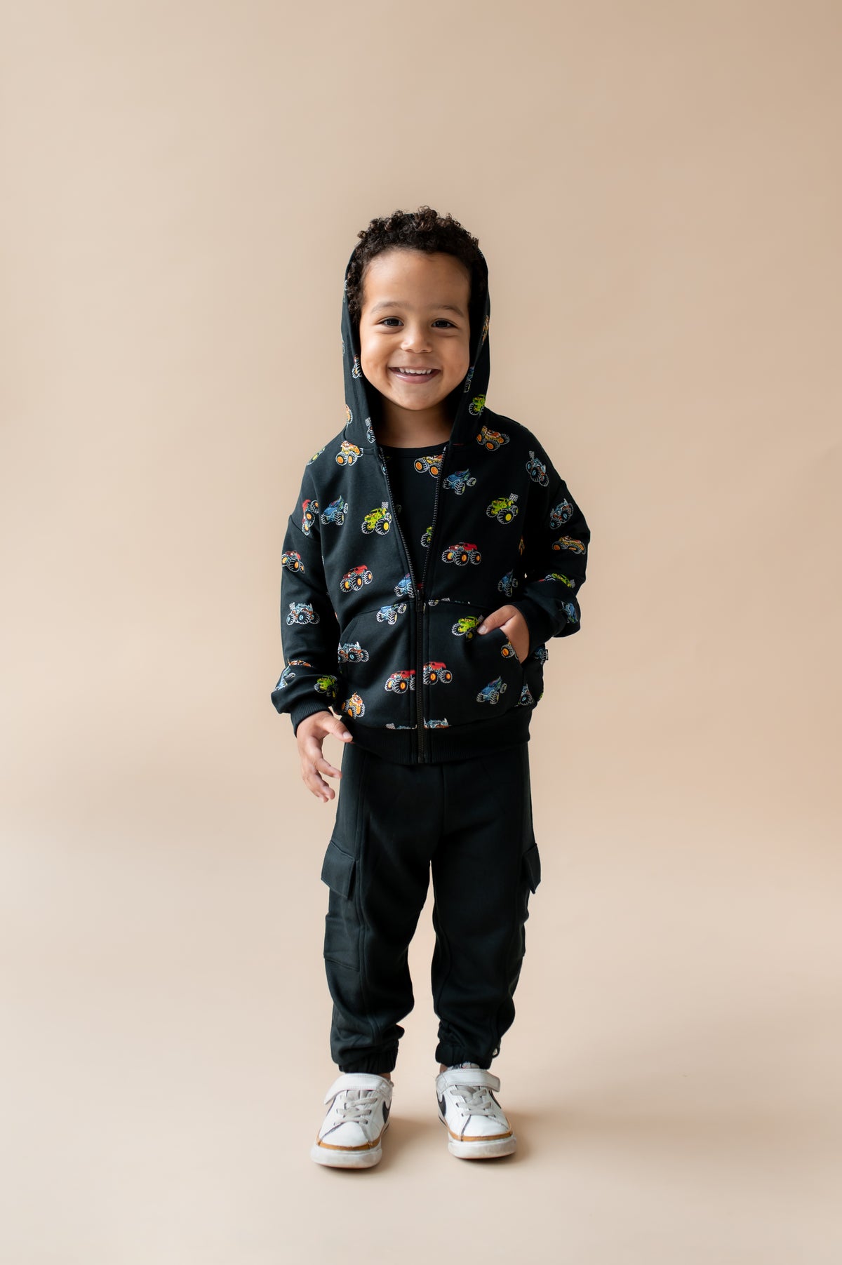 Boy standing wearing Black Zip-Up Hoodie in Monster Truck print over black pant and white shoes on a beige background