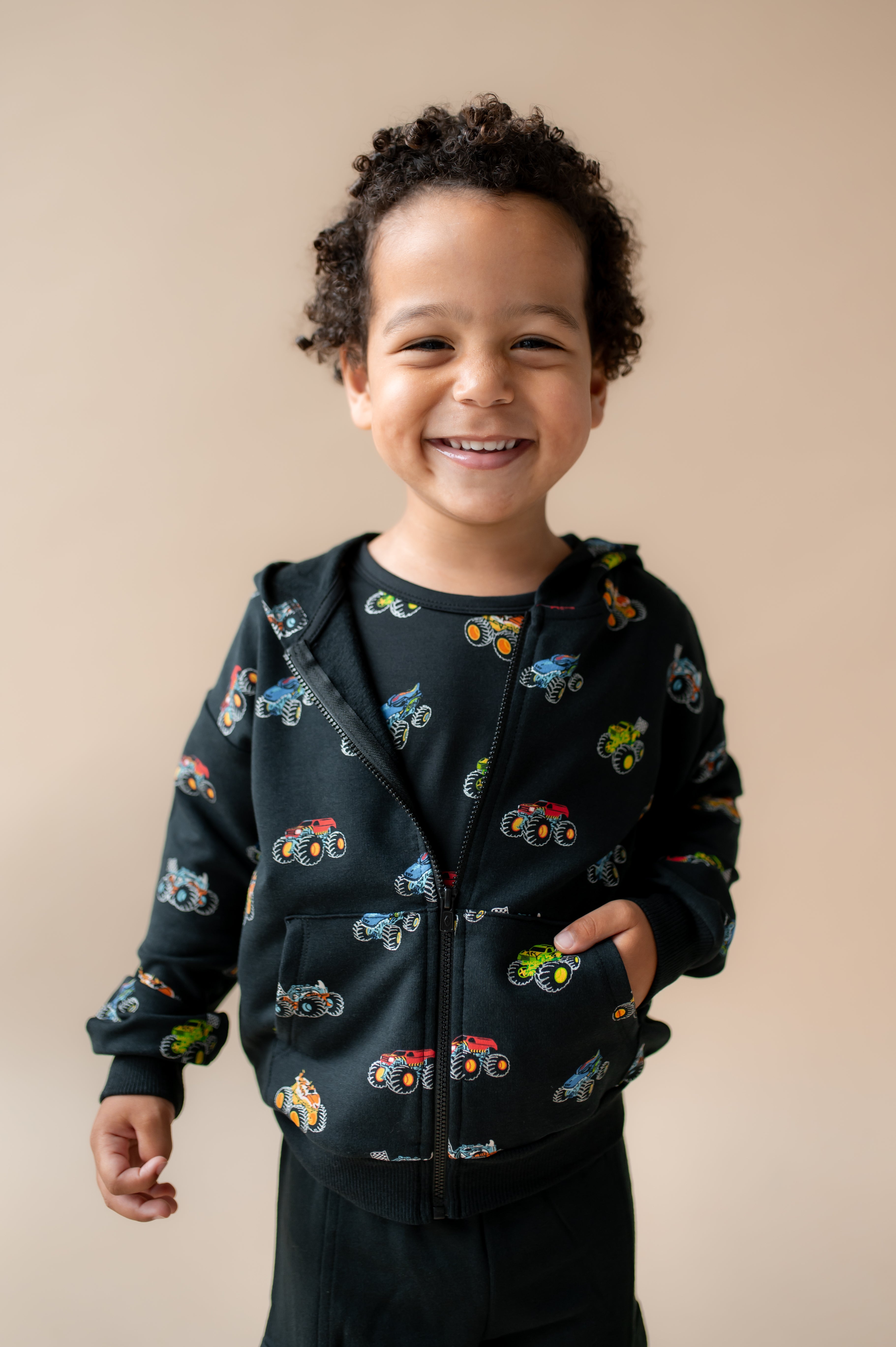 Boy smiling in Black Zip-Up Hoodie in Monster Truck print and black pant on a beige background